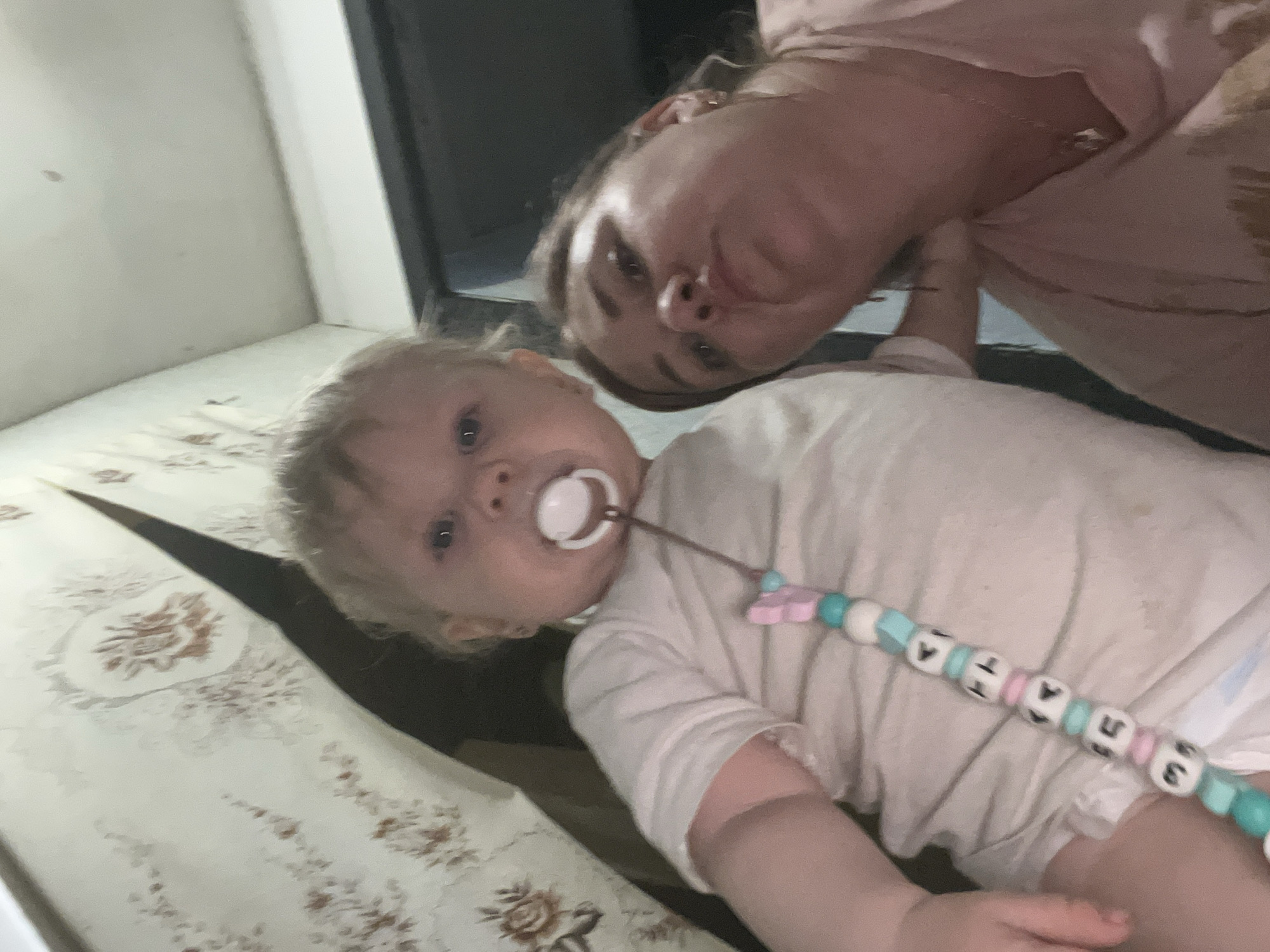 In a dimly lit interior space, possibly a residential room in the city of Fokino, Russia, a woman and a baby are captured in a low-angle selfie. The woman, with blonde hair pulled back, is in the foreground, her face partially obscured by the baby and her own arm. She is wearing a pinkish-toned garment. To her left, a baby with light hair and a pacifier in its mouth is the primary focus. The baby is wearing a white t-shirt and appears to be leaning forward. A beaded pacifier clip, adorned with letters and colorful beads, dangles from the pacifier. The background shows a wall with patterned wallpaper and a doorway or opening revealing a dark space. The lighting is uneven, suggesting it might be nighttime or the interior is poorly lit. The overall scene is informal and appears to be a spontaneous moment captured by the woman.
