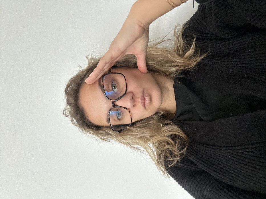 A female subject is depicted in a close-up, chest-up portrait against a plain white background, consistent with an indoor location in Veauchette, France. The subject has light brown, wavy hair and is wearing large, square, tortoiseshell-patterned eyeglasses. Her left eye appears blue, and her right eye appears green-hazel. Her lips are slightly pursed. She is wearing a black ribbed cardigan over a dark top, with a thin gold chain visible around her neck. Her left hand is raised to her forehead, fingers extended towards her temple, forming a 'salute-like' or contemplative gesture. A delicate beaded bracelet is visible on her right wrist.