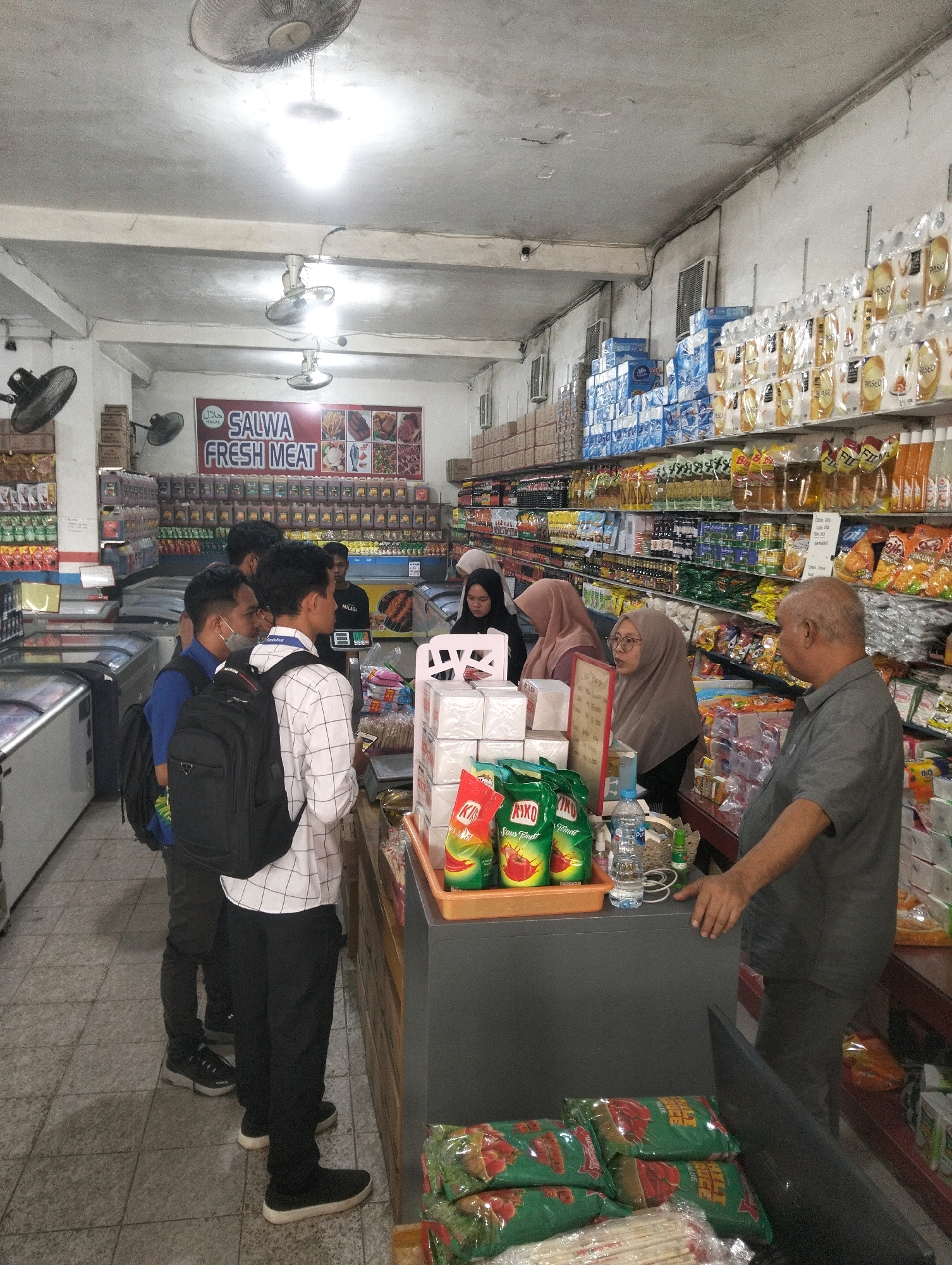 In the city of Palangka Raya, Indonesia, a grocery store is depicted. The store appears to be a small to medium-sized establishment with aisles of products, refrigerators, and a checkout counter. The lighting is fluorescent and slightly dim, casting shadows.
Several individuals are present in the store. At the checkout counter, an older man in a grey shirt is interacting with a younger woman wearing glasses and a headscarf. Another woman in a dark hijab is also visible near the counter. In the foreground, three young men are standing together. One is wearing a blue shirt and a black backpack, another has a white and black checkered shirt and a black backpack, and the third is wearing a plaid shirt and a black backpack. A fourth man, wearing a dark shirt, is standing behind the checkout counter.
The store is stocked with various goods. Shelves are filled with bottled drinks, packaged food items like snacks and sauces, and what appear to be cleaning supplies. A prominent sign with the text "SALWA FRESH MEAT" is displayed, along with images of various meat products. Refrigerated display cases are also visible, some of which are open. The floor is tiled. The overall impression is that of a busy local market.