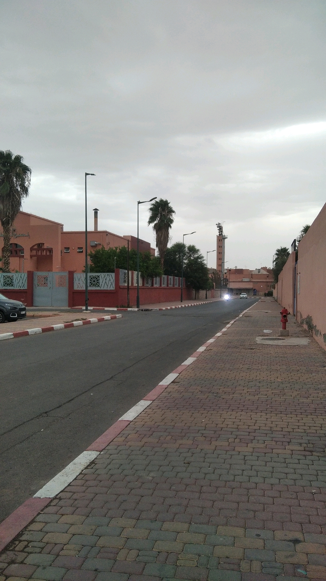A straight, paved road with a red and white striped curb runs through a city. The sky is overcast. On the left, there is a pink building with a fence in front of it, and a palm tree. Further down the road on the left, there are more buildings and trees. On the right side of the road, there is a cobblestone sidewalk, and a red fire hydrant. In the distance, a tall, narrow building with a flat top and what appears to be lights on it, stands against the sky. A white car is driving down the road towards the tall building.