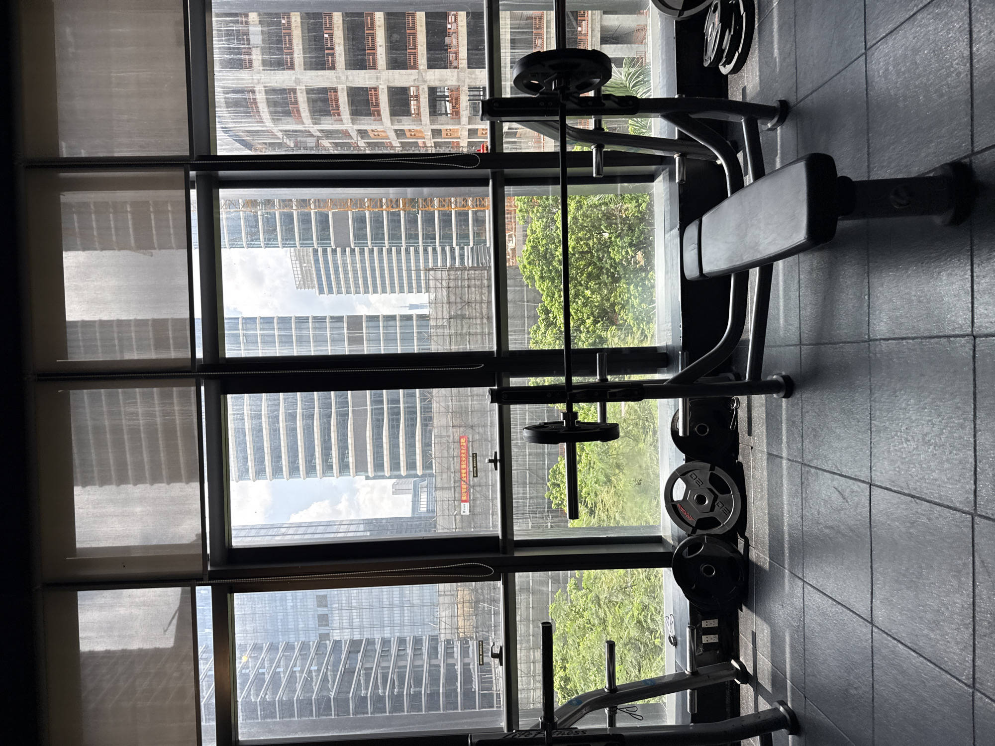 The image depicts an indoor gym with a weight bench and barbell in the foreground. In the background, a large window offers a view of a city skyline, featuring modern skyscrapers and ongoing construction. The scene is well-lit by natural light filtering through the window. The gym floor is covered in black rubber tiles. On the left side of the image, a weight rack is partially visible. Several weight plates are placed on the floor near the bench. The view through the window suggests an urban environment, likely in a developed area of Haizhu District, China, characterized by high-rise buildings and visible construction cranes. There are no people present in the image.