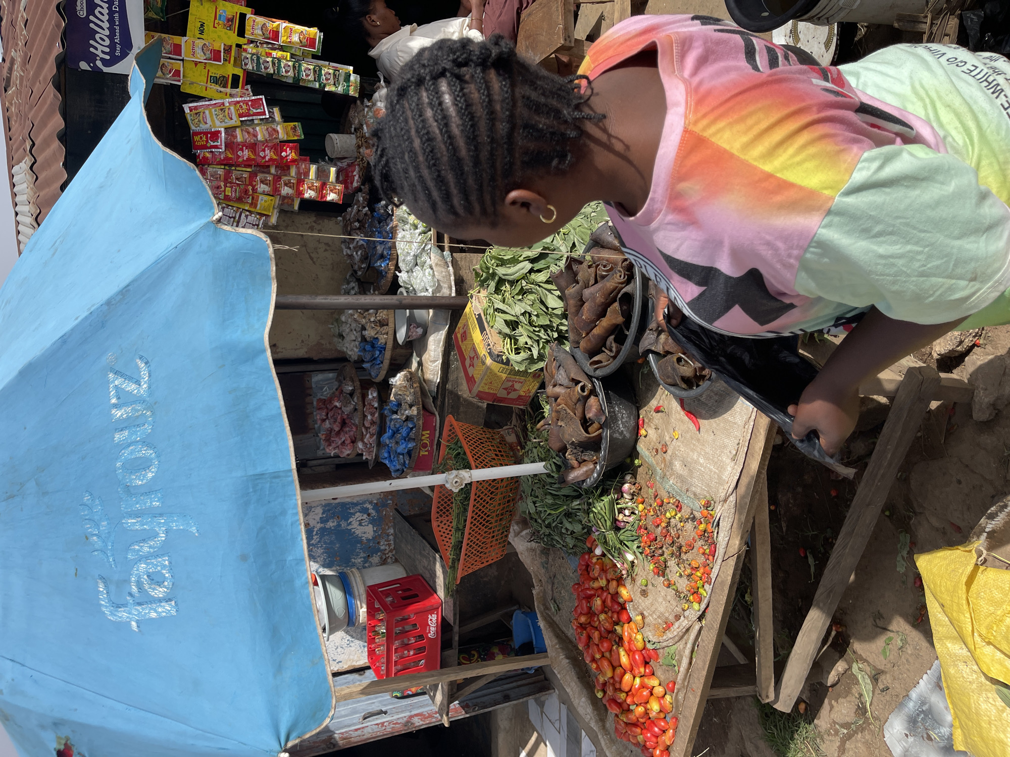 A woman is selecting produce at a roadside market stall in Jos, Nigeria. The stall features a variety of goods, including tomatoes, greens, and dried items in buckets. Packaged goods hang overhead, and a Fayrouz branded umbrella provides shade. A Coca-Cola crate is visible below the counter.
