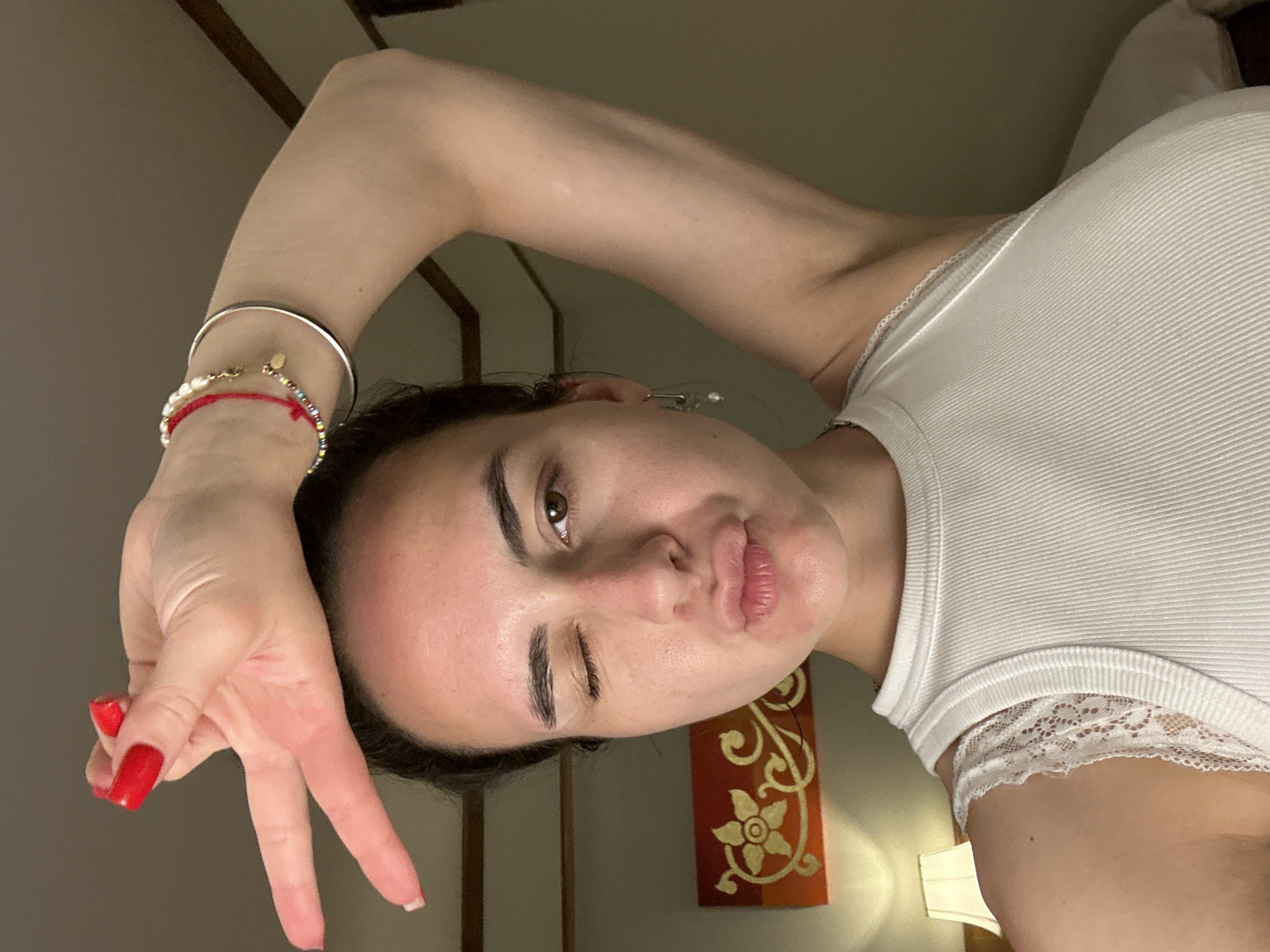 A young woman with fair skin and dark hair is positioned at an oblique angle facing the camera, winking her left eye. She is wearing a white ribbed tank top with lace trim around the neckline. Her left arm is raised, with her hand making a peace sign near her forehead. She has multiple bracelets on her left wrist. Her nails are painted red. Her lips are pursed.
The background is a dimly lit room. A decorative panel with a floral pattern is visible on the wall behind her. The location is presumed to be Pattaya City, Thailand, but there are no immediately obvious landmarks to confirm the location.