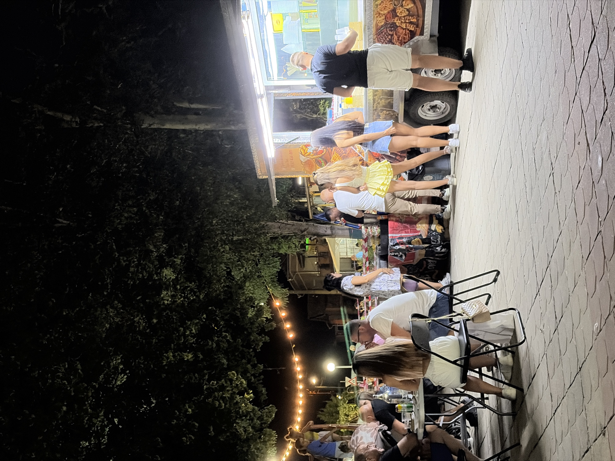 The image depicts an outdoor scene at night, possibly in the city of Rupite, Bulgaria, with a food truck as a central focal point. Several people are gathered, some standing in line at the food truck, while others are seated at tables. String lights are visible overhead, illuminating the area. The ground is paved with interlocking stones. The food truck has an illuminated serving window and displays images of food on its exterior. The overall atmosphere suggests a casual social gathering or event.