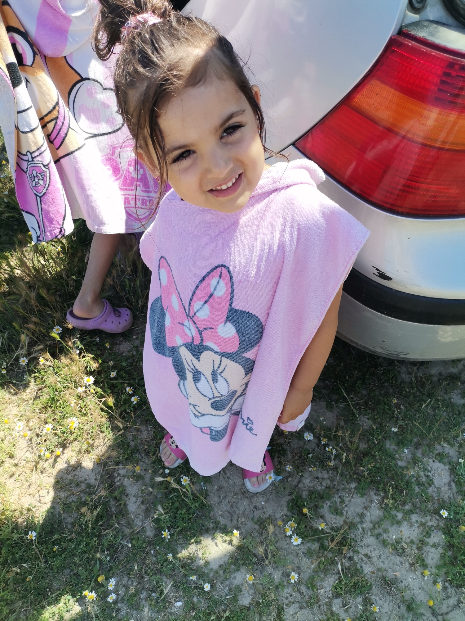 A young girl, wearing a pink poncho with a Minnie Mouse design and pink sandals, stands next to a car. Her hair is pulled back in a ponytail. She is smiling at the camera. The ground is covered in dry grass and small white flowers. A car's red taillight is visible in the background. The image appears to have been taken outdoors in a sunny environment, possibly in Kranevo, Bulgaria.