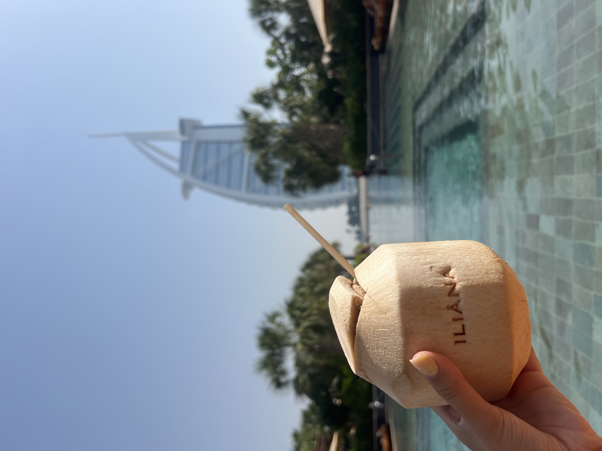 A human hand holds a fresh coconut drink, engraved with the name "ILIÁNA", with a straw inserted, against a backdrop featuring the Burj Al Arab Jumeirah hotel in Dubai, United Arab Emirates. The hotel is partially visible in the background, with a clear blue sky above it. The foreground includes a pool with light blue tile, while trees and vegetation are interspersed between the drink and the hotel, slightly blurring the scene.