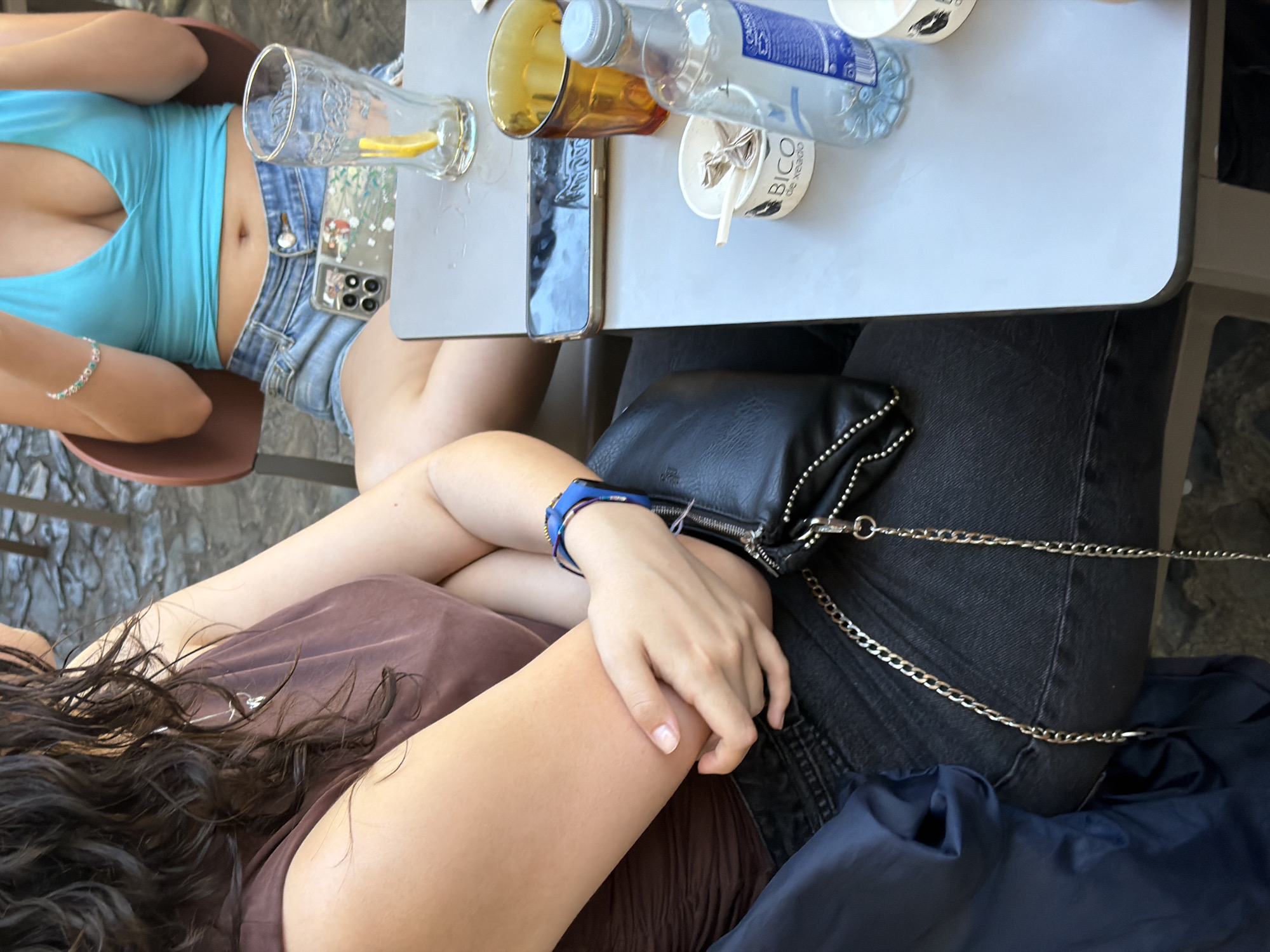 The image depicts two individuals seated at an outdoor table. The person in the foreground, on the left, has long, dark, curly hair and is wearing a brown top and black jeans. They are holding their arm across their chest, and a black purse with a silver chain strap is visible. On the right, another person is wearing a turquoise halter top and denim shorts. They are holding a smartphone with a decorative case. On the table, there are two drinking glasses, one containing a slice of lemon and the other amber-colored. A bottle of water and a small container with a spoon are also present. The background shows a cobblestone surface, likely a street or patio, and a portion of a chair. The setting suggests a casual outdoor dining or cafe environment in Santiago de Compostela, Spain.