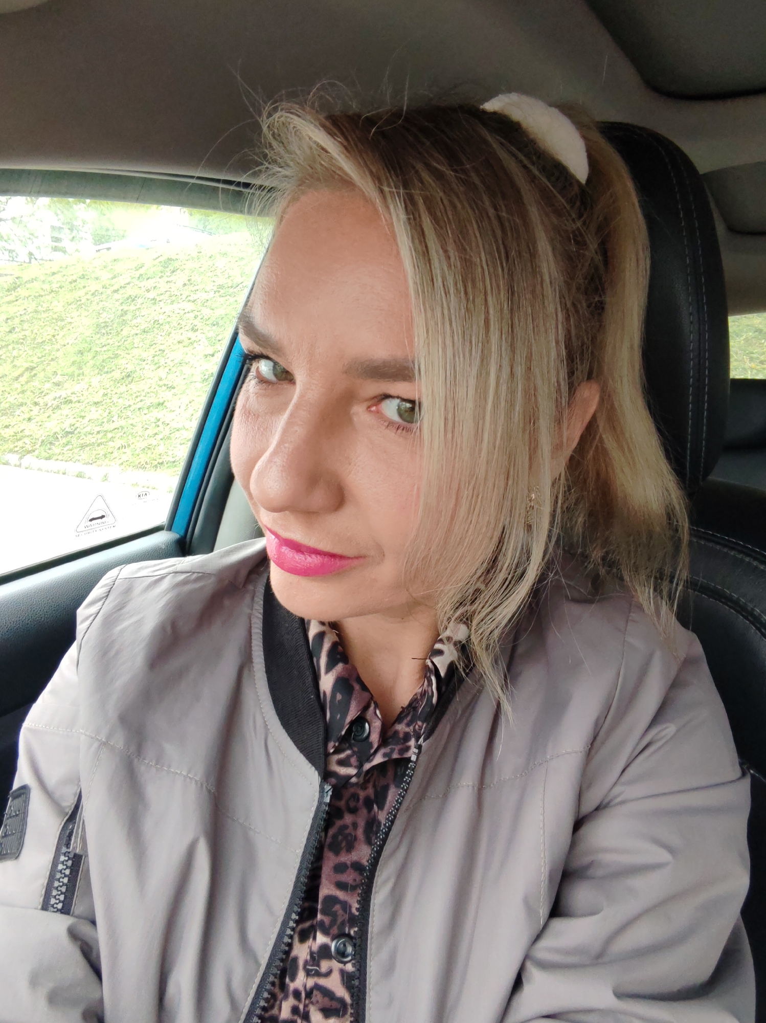 A woman with blonde hair pulled back in a ponytail with a white scrunchie is in the driver's seat of a car. She is wearing a gray bomber jacket over a leopard print shirt. Her makeup includes bright pink lipstick. She is looking at the camera with a slight smile. The car's interior is visible, including the black leather seat and the gray headliner. Outside the car window, a green, grassy hill is visible. The image appears to be a selfie taken by the woman in the car.