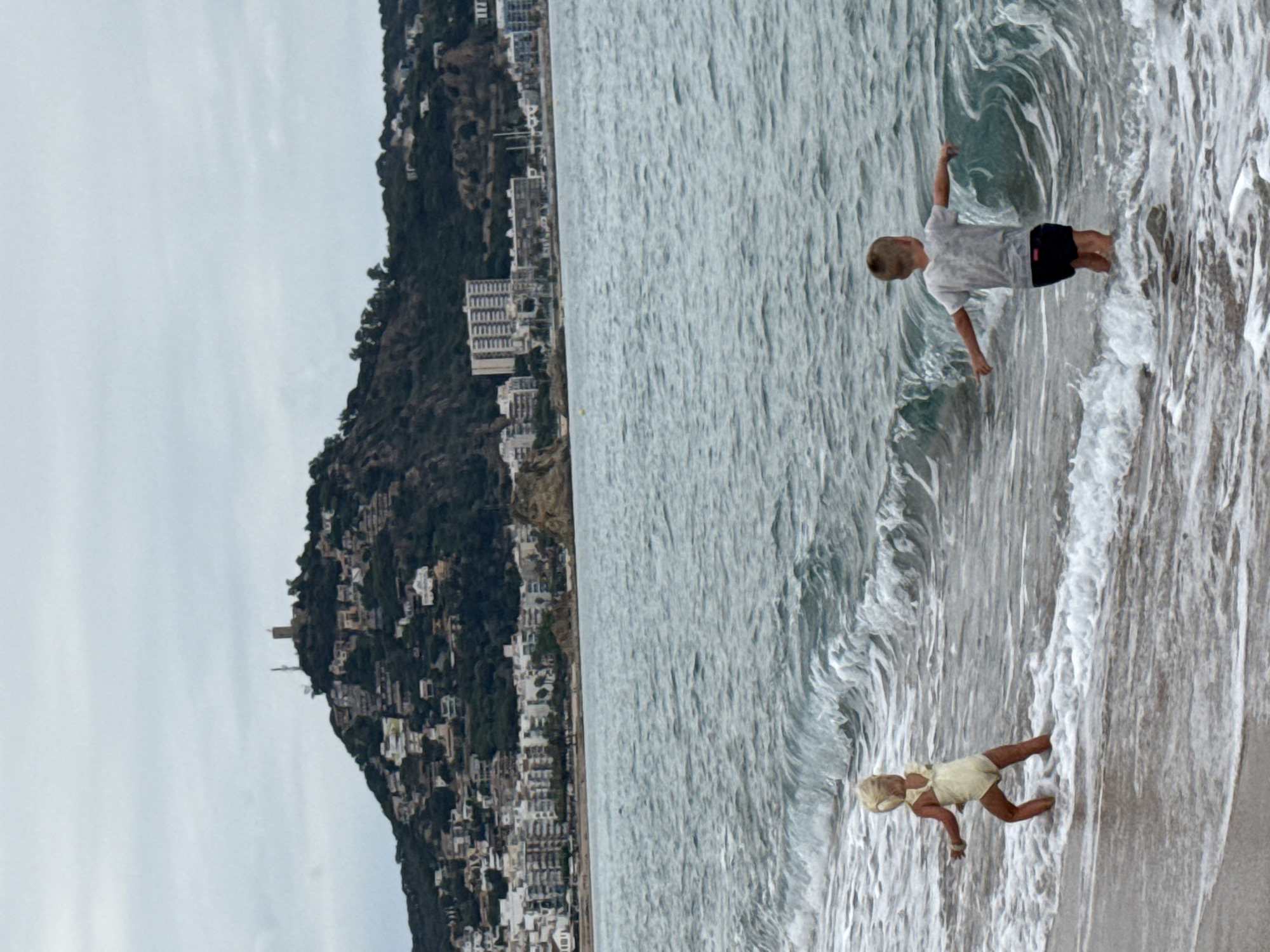 In the foreground, two young children, a girl and a boy, stand in the shallow water at the edge of the sea. The girl, wearing a yellow swimsuit and a bonnet, is positioned on the left, facing away from the camera and towards the sea. The boy, on the right, is also facing away from the camera, dressed in a grey t-shirt and dark shorts, with his arms outstretched to his sides. Waves are breaking around them, creating white foam.
In the midground, the calm sea extends towards a coastline dotted with buildings.
The background features a prominent hill covered in dense green vegetation and a scattering of buildings. At the crest of the hill, a stone tower stands, a historical landmark. The town of Blanes, Spain, is visible spread across the lower slopes of the hill and along the coastline, characterized by a mix of modern and older architecture, including apartment buildings and smaller houses. The sky above is overcast, with muted grey clouds. The overall impression is a coastal scene in Blanes, Spain, with the town and its natural surroundings under a cloudy sky, and two children enjoying the water.
