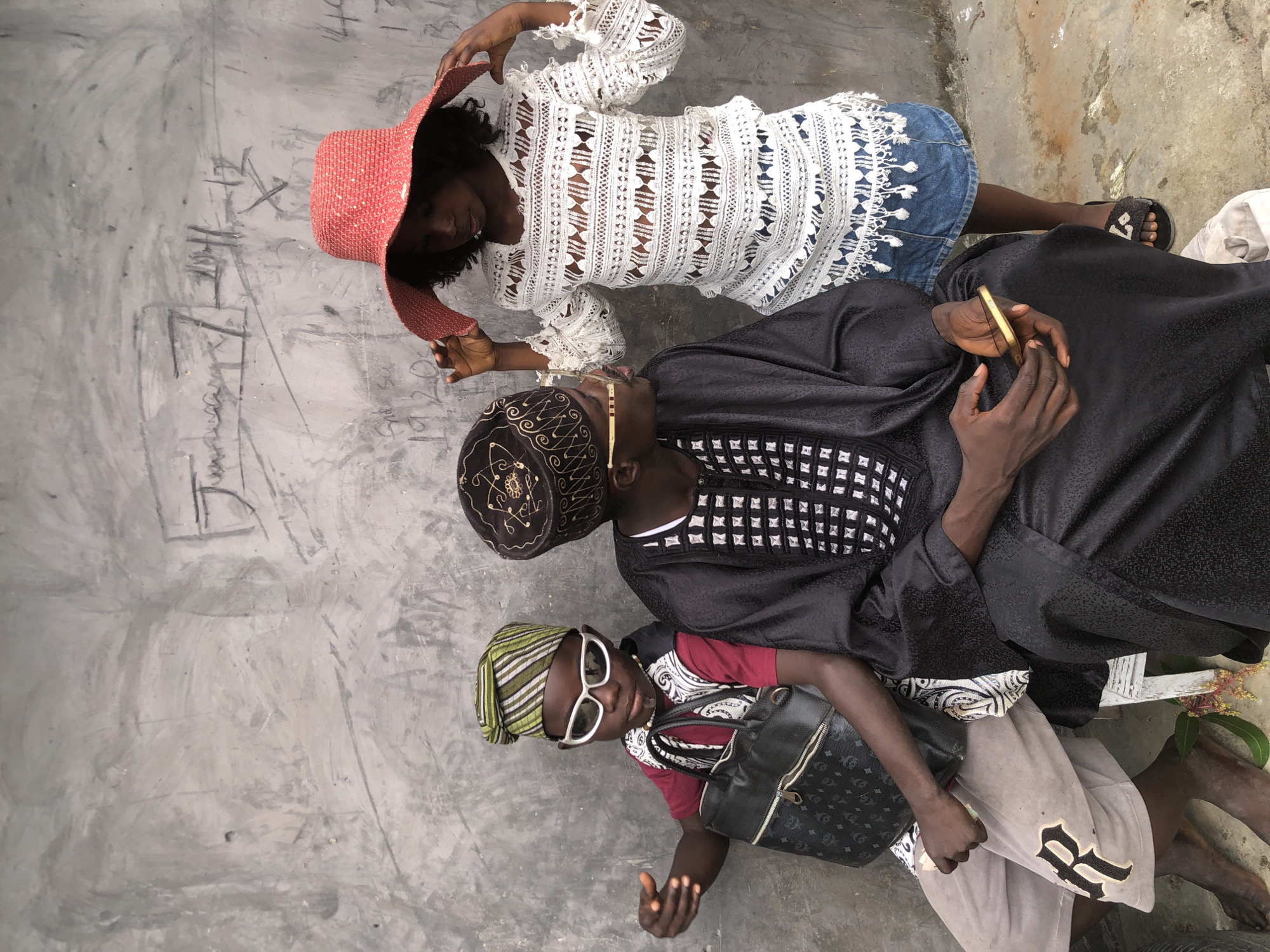 In Ado, Nigeria, three individuals are posed in front of a rough, grey concrete wall that is graffitied with what appears to be chalk markings, including dates and illegible symbols.
On the left, a young person, likely a child, is seated and wears a maroon t-shirt layered with a white vest patterned with black accents. They sport a green and white striped headwrap and white-rimmed sunglasses. The child holds a black handbag with a gold emblem and chain strap. Their legs are crossed, with one foot showing a portion of light-colored shorts with a large, dark 'R' logo.
In the center, an adult, likely male, is seated and dressed in a black, traditional garment adorned with a white, patterned collar and intricate black embroidery on the chest. They wear a dark, embroidered cap. The individual is looking to their right, towards the person next to them, and holds a gold-colored mobile phone in their hands, which are resting on their lap.
On the right, a young person, likely a girl, is standing and wearing a white, lacy top with an open-weave pattern and frayed edges. Below the top, a denim skirt is visible. They are wearing a wide-brimmed, red hat adorned with a subtle pattern and are touching the brim with both hands. The girl is looking down and slightly to her left. She wears black sandals with a decorative embellishment. The three subjects are positioned closely together, suggesting a familial or social connection. The ground is concrete, with some scattered debris and a small green plant with reddish flowers visible near the seated individuals' feet.