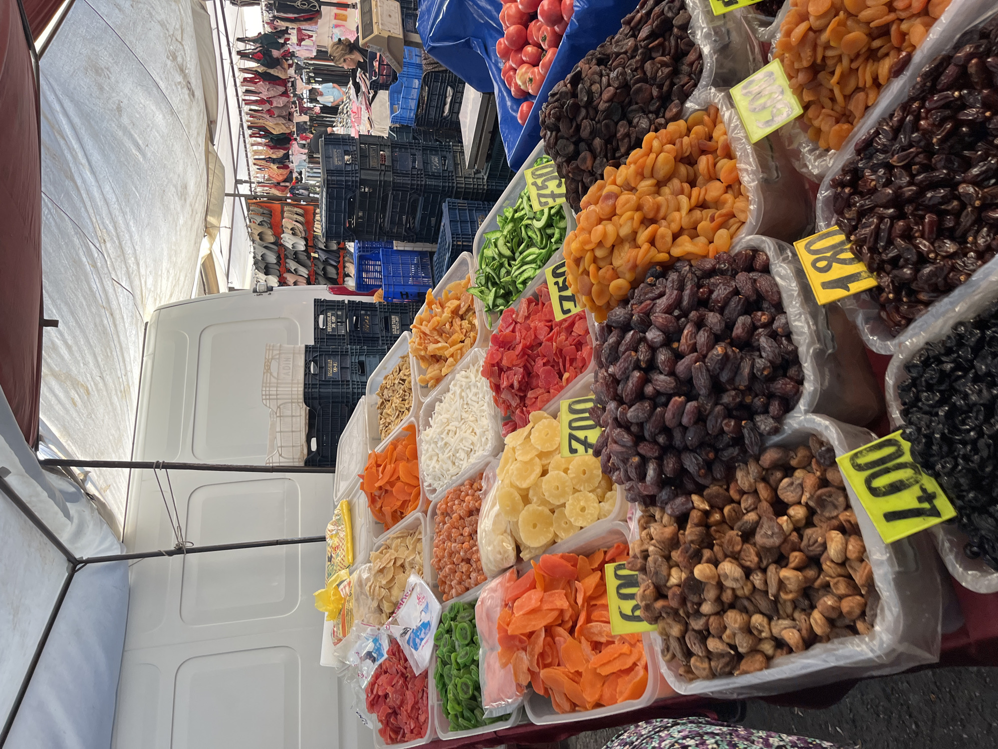 This is a street market scene in Alanya, Turkey, showcasing a vibrant display of dried fruits and other food items. The foreground is dominated by several large, transparent plastic containers filled with a variety of colorful dried fruits. These include dark brown dates, lighter brown figs, bright orange dried apricots, green dried fruit slices, and red dried fruit pieces. Some containers have yellow price tags with numbers printed on them, such as "600", "700", and "750". Behind these, further containers hold more dried fruits and what appear to be dried apricots. To the right, a large blue tarp covers a collection of red tomatoes.
In the background, the market extends into the distance. Several dark plastic crates are stacked behind the food stalls, and further back, rows of women's footwear, handbags, and clothing are displayed on racks, suggesting a diverse market offering. A white van is parked behind the stall, and a portion of its white side is visible. Two individuals are visible in the background: a woman with blonde hair is standing, and another person in a light blue shirt is seated. The scene is set under a white and maroon striped canopy, indicating an outdoor market. The overall impression is one of a bustling marketplace filled with fresh and preserved produce and goods.