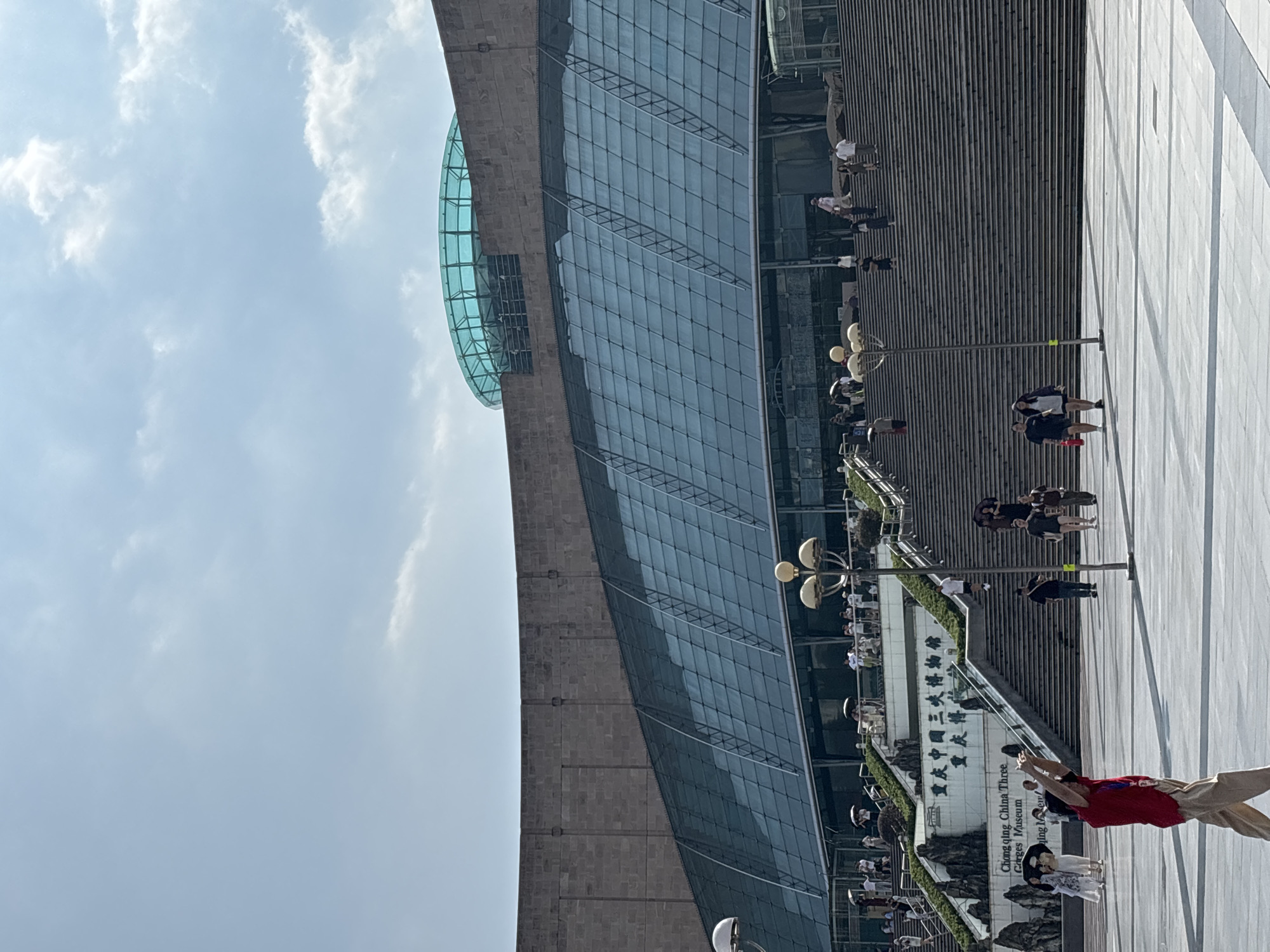 The image depicts the Chongqing China Three Gorges Museum, located in Yuzhong District, China. The museum's facade features a large, curved glass-paneled structure extending upwards, topped by a distinctive turquoise-colored circular glass roof. The building is set against a backdrop of a bright, partly cloudy sky. A grand, wide staircase leads up to the museum's entrance, with numerous people visible ascending and descending. Some individuals are holding umbrellas, suggesting sunny weather. In the foreground, a person in a red shirt and beige pants is captured in mid-motion, possibly posing for a photo or performing. Other individuals are scattered across the plaza and on the stairs, engaging in casual movement and conversation. The plaza in front of the museum is paved with large, light-colored square tiles. The overall scene captures a lively public space with people visiting a prominent cultural institution on a clear day.