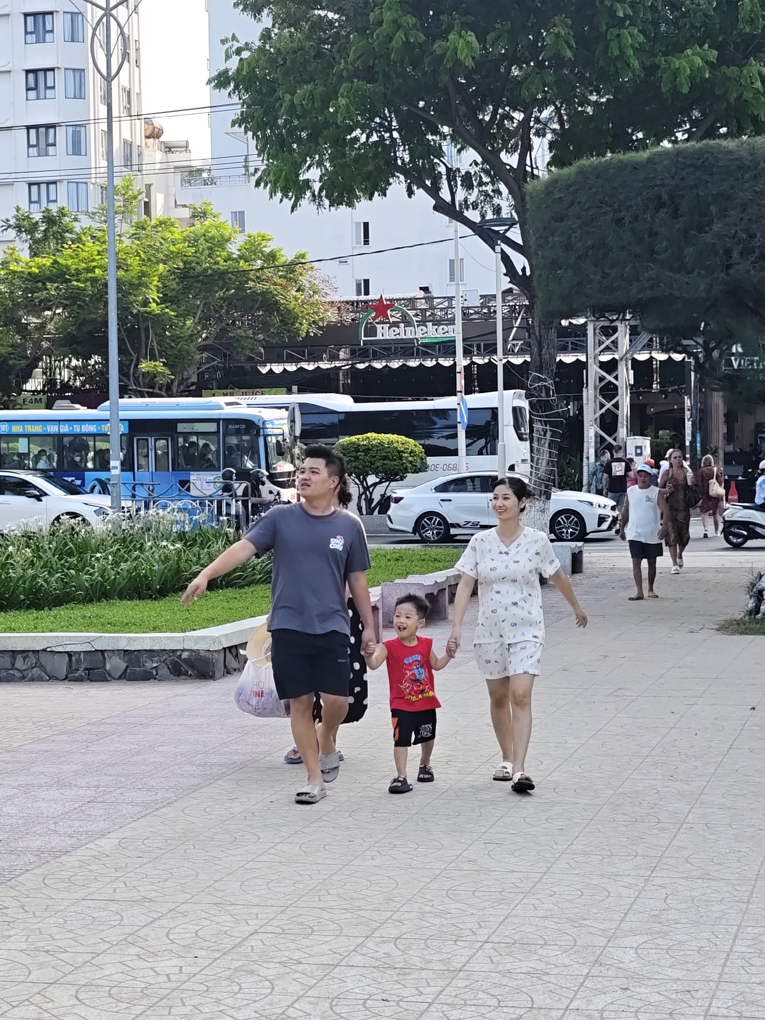 In Nha Trang Ward, Vietnam, a family of three is walking on a paved pedestrian pathway lined with grass and low stone walls. The father, on the left, wears a grey t-shirt and black shorts, carrying a bag in his left hand. Beside him, a woman in a white patterned outfit, likely the mother, walks hand-in-hand with their young son. The son, wearing a red shirt with "Spider-Man" printed on it and black shorts, is between them. In the background, a blue and white bus is visible on a street, along with other pedestrians, a white car, trees, and buildings. The scene captures a moment of family leisure in an urban outdoor setting.