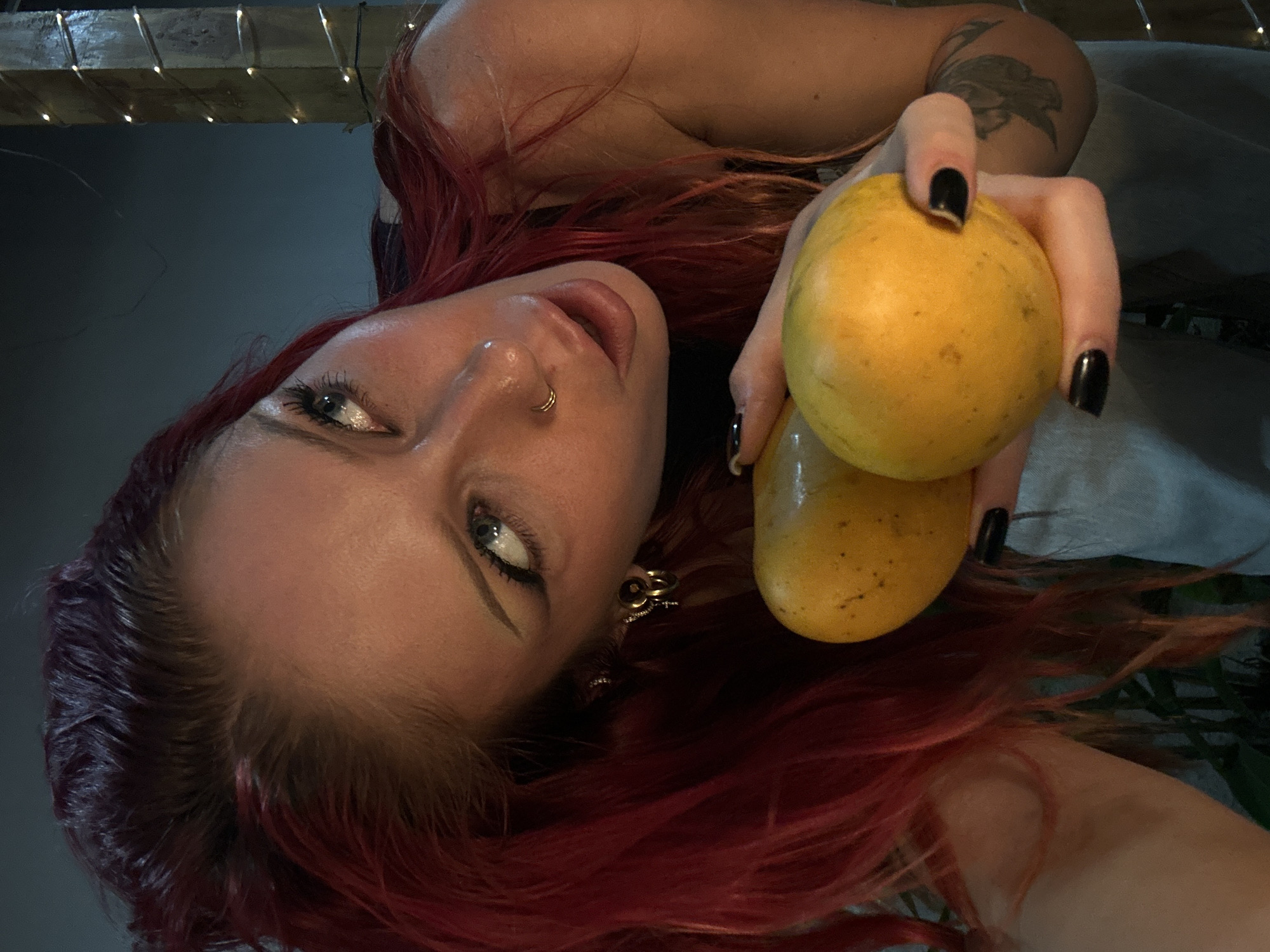 A woman with vibrant red hair holds two yellow fruits, possibly mangoes or papayas, in her right hand. She is leaning in towards the camera, looking upwards and to her right with wide eyes. Her mouth is slightly open, and she has a nose ring and earrings. She is wearing a dark top and light-colored clothing around her waist. The background is dark and out of focus, with some faint lights visible on a wooden structure on the right side of the frame, suggesting an outdoor setting at night. The woman has visible tattoos on her arm. The image appears to be taken in North Kuta, Indonesia.