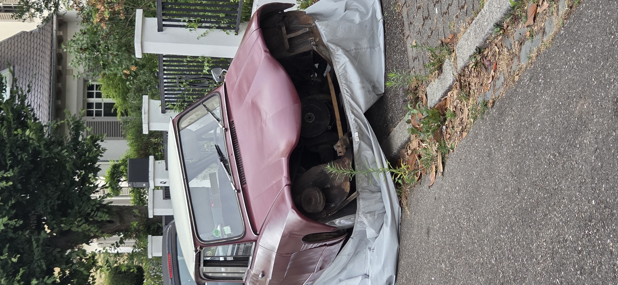 The image shows a severely damaged, maroon-colored vintage car, possibly a 1960s model, parked on a street. The front of the car is missing, revealing the engine and internal components. A light gray car cover is draped over the damaged front section and extends downwards, partially obscuring the wheels and the ground. The car is parked next to a sidewalk with cobblestone and paved sections, bordered by a curb. Several small green plants and dry leaves are visible growing around the base of the car and along the sidewalk. In the background, a dark metal fence with white posts stands behind a black mailbox on a pedestal. Beyond the fence, a building with a dark tiled roof and shuttered windows is visible, partially obscured by the branches of a large, leafy tree. The overall scene suggests the car is either abandoned or undergoing extensive repair. The location is a residential street in Strasbourg, France, characterized by typical European architecture and landscaping.