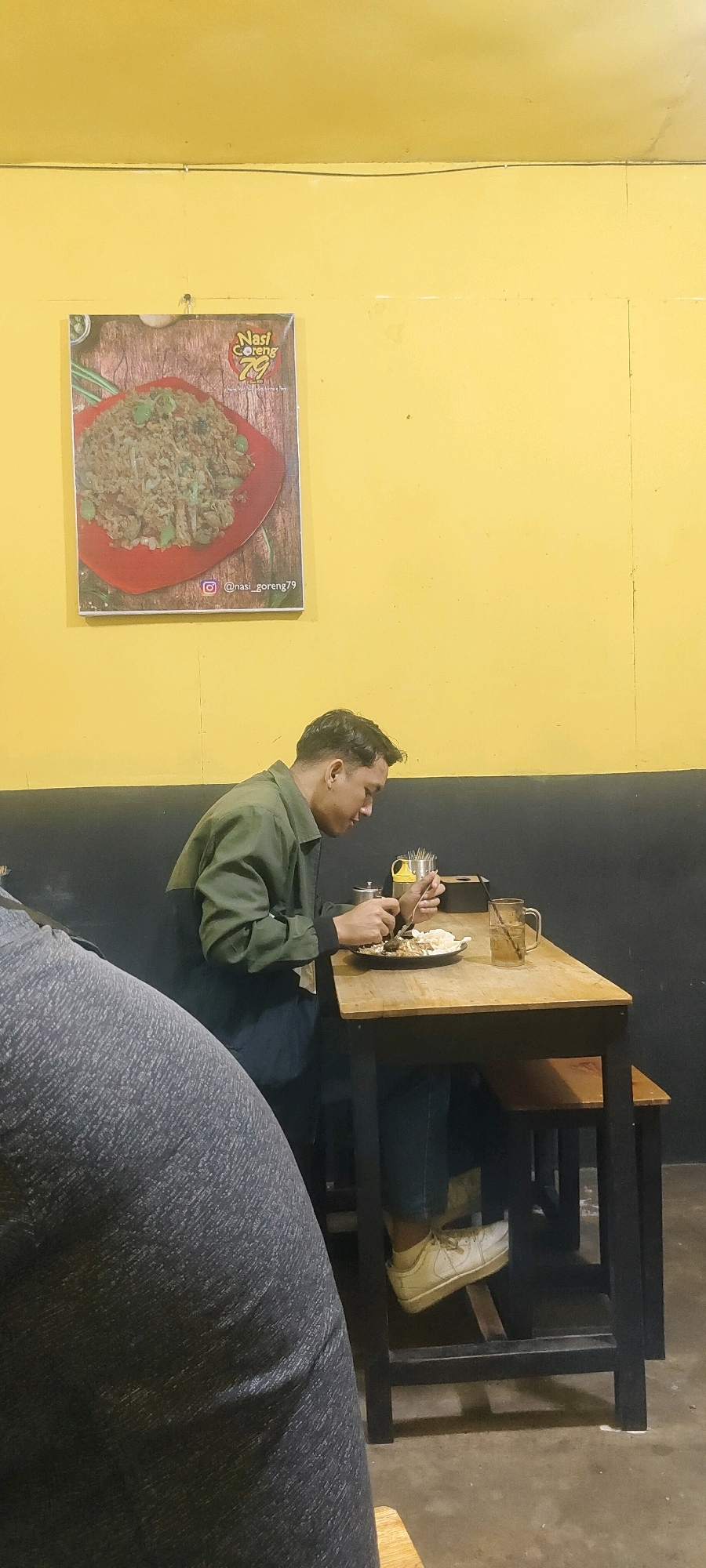 A man, appearing to be of Southeast Asian descent, is seated at a wooden table in what appears to be a restaurant. He is wearing a dark green jacket over a lighter shirt and dark pants, with white sneakers. He is holding a fork and knife, in the process of eating from a plate of food. A glass of amber liquid, likely tea or water, sits on the table. A poster depicting a plate of nasi goreng is hanging on a yellow wall behind him, with the handle "@nasi_goreng79" visible. The restaurant has a dark-colored lower wall. The flooring appears to be concrete. The overall lighting suggests an indoor setting, possibly in the late afternoon or evening. The image was taken in Palangka Raya, Indonesia.