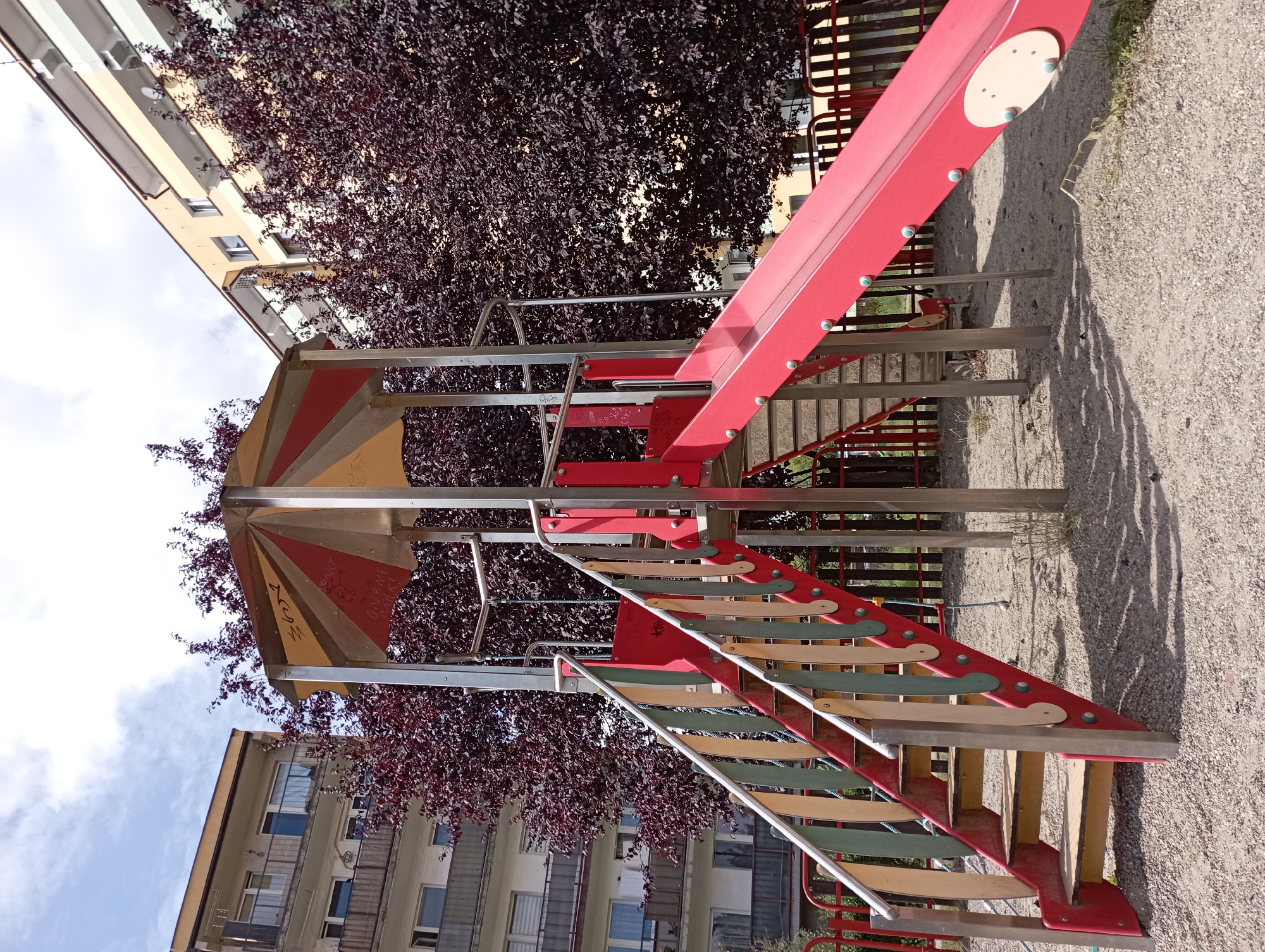 The image depicts a children's playground structure in Siemianowice Śląskie, Poland, characterized by a multi-level play tower with a red slide, a staircase with colorful wooden railings, and a covered top section. The playground is situated in an urban setting, with apartment buildings visible in the background. The buildings are multi-story structures with balconies and windows, suggesting a residential area. Dark red, leafy trees surround the playground, creating a dense canopy. The ground is covered with gravel and sand, typical of playground surfaces. The sky is partly cloudy, indicating daylight. There are no people present in the image. The overall scene suggests a public recreational space within a city.