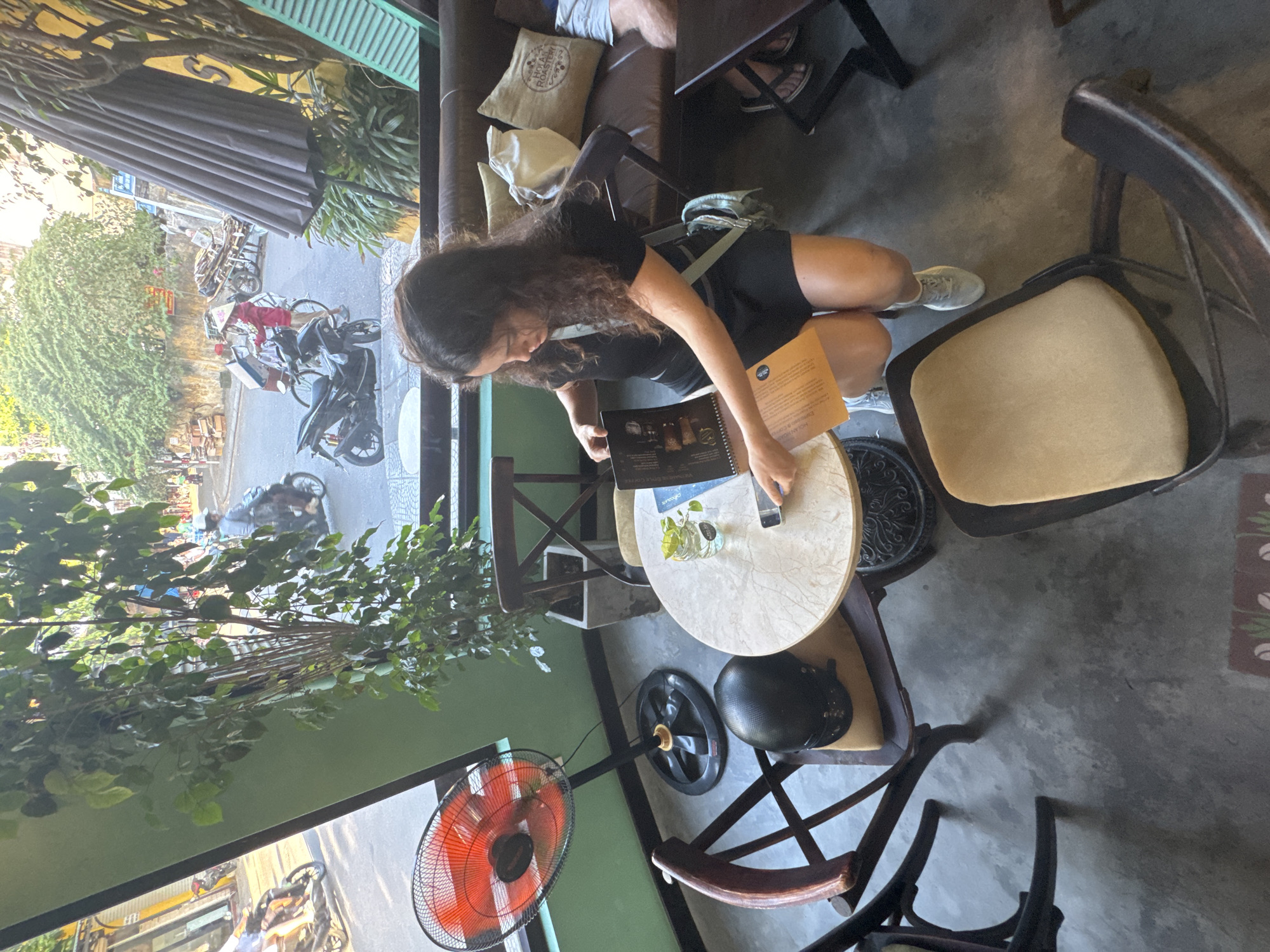 A young woman with curly brown hair is sitting at a round cafe table, looking at a menu. She is wearing a black t-shirt and black shorts, with white sneakers. A light blue crossbody bag is slung over her shoulder. On the table, there is a glass of water with a sprig of mint, a mobile phone, and a menu. A black helmet rests on a chair next to her. In the background, a brown leather sofa is visible with decorative pillows. The cafe appears to be open-air, with a view of a street in Hoi An, Vietnam, featuring motorbikes and pedestrians. A large green plant is in the corner of the cafe. A red oscillating fan is on the floor to the left.