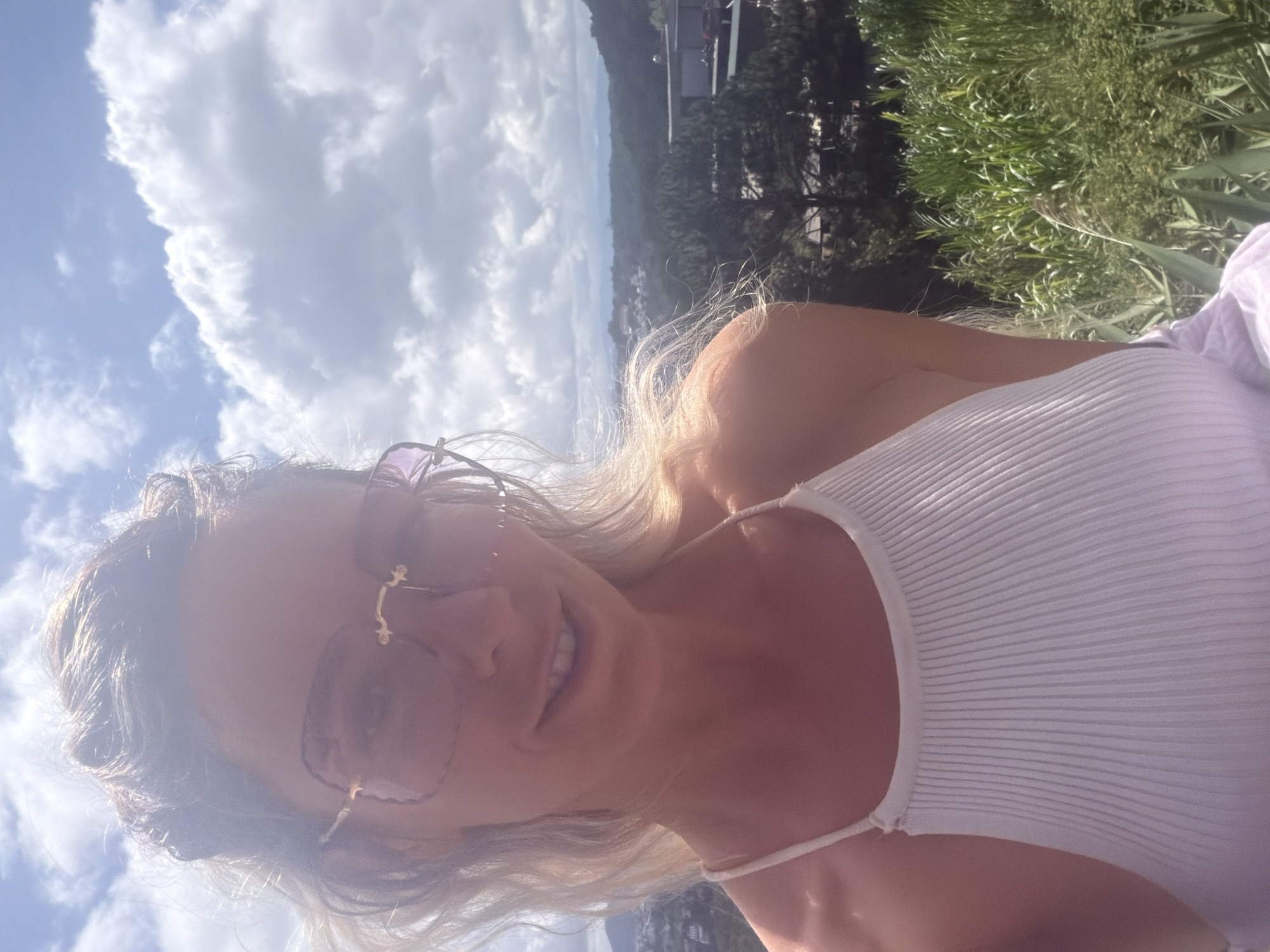 A close-up, selfie-style shot features a woman with blonde hair and large, pink-tinted sunglasses. She is wearing a white ribbed tank top and is smiling at the camera. Her hair is partially obscuring her face, and the bright sunlight creates a glare effect. The background shows a lush, green landscape with trees and buildings, indicative of the city of Xuân Trường - Đà Lạt, Vietnam. The sky above is filled with scattered clouds.