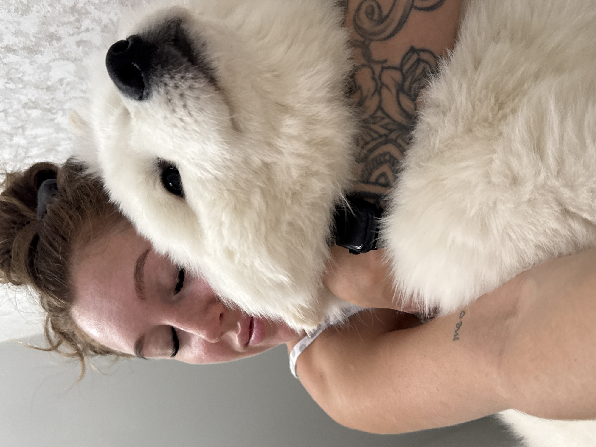 The image captures an intimate moment between a woman and a fluffy white dog in what appears to be an indoor setting. The woman is holding or cuddling the dog, whose head is resting affectionately against her. Her eyes are gently closed or looking downwards, conveying a sense of peace and tenderness. Her medium-brown hair is styled loosely, and she is wearing a light-colored top, possibly a tank top or camisole.
Notable details on the woman include a large, intricate tattoo of floral and mandala-like patterns on her right arm and shoulder area. On her left forearm, a small, subtle tattoo spells "me ♡". The dog, likely a Samoyed due to its dense, pristine white fur, black nose, and dark, expressive eyes, appears calm and content, nestled against the woman. It wears a simple black collar.
The background is simple, featuring a light-colored, textured surface on one side (possibly a blanket or a soft wall) and a plain light gray surface on the other, suggesting a cozy indoor environment. The lighting is soft and diffused, indicative of daytime. The overall emotion conveyed is one of deep affection and warmth. While the location context of Chaikovsky, Russia, is provided, there are no specific visual cues within the image itself to confirm this geographical detail.