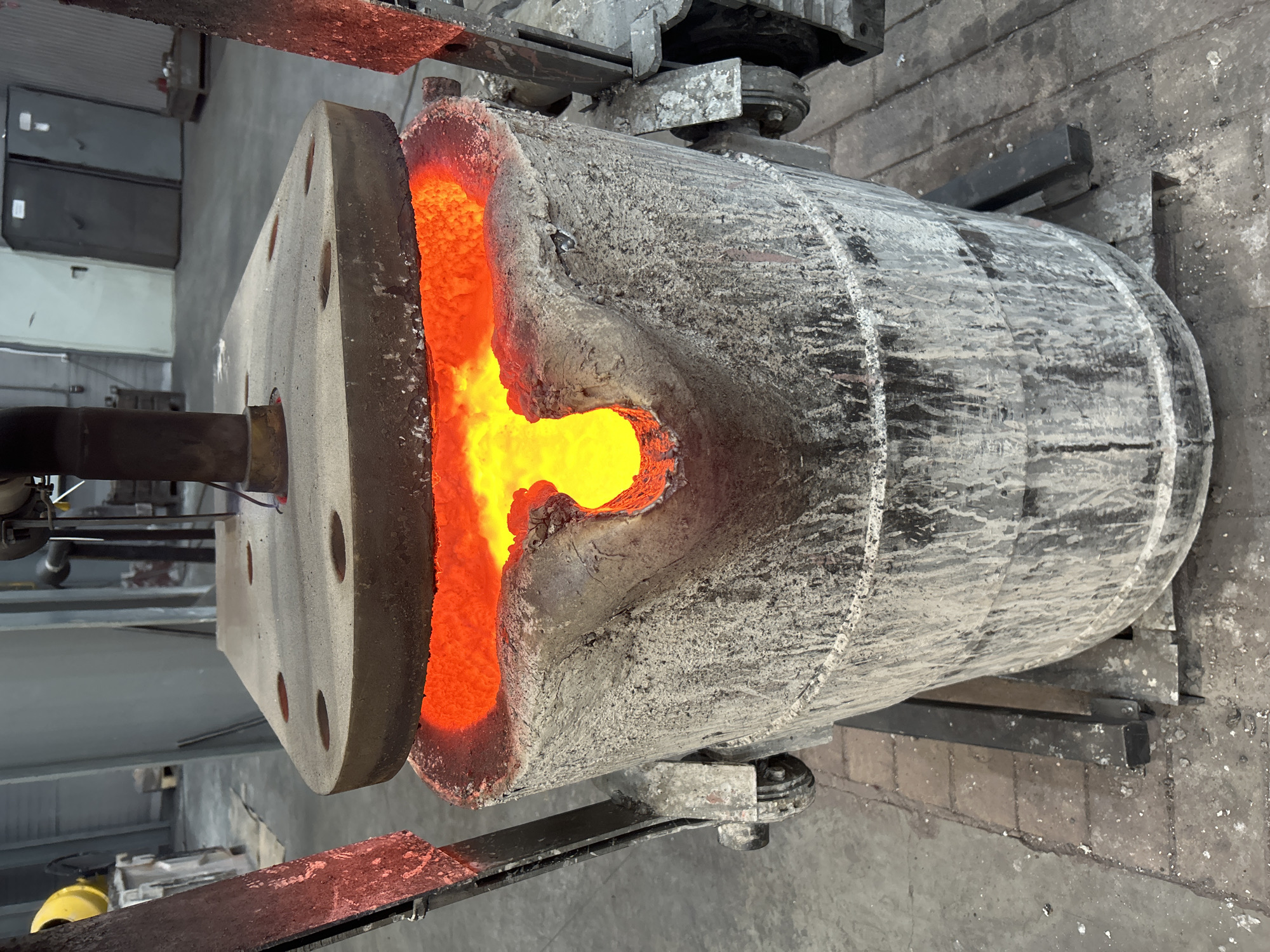 The image depicts a metallurgical process, most likely inside an industrial facility, but the city of Noviy, Russia, cannot be verified as the location. A large, cylindrical furnace is suspended by a mechanical rig above a concrete floor. The furnace appears to be made of a refractory material coated with a dusty, gray-white residue. The interior of the furnace is visible through an opening, glowing intensely orange and yellow due to the high heat. Molten material appears to be visible in the opening. A thick, flat flange is attached to the furnace, with a cylindrical structure projecting from its center. The flange has multiple circular holes drilled around its perimeter. The surrounding environment is industrial, with the metal framework of the support structure visible. The floor shows signs of wear and tear.
