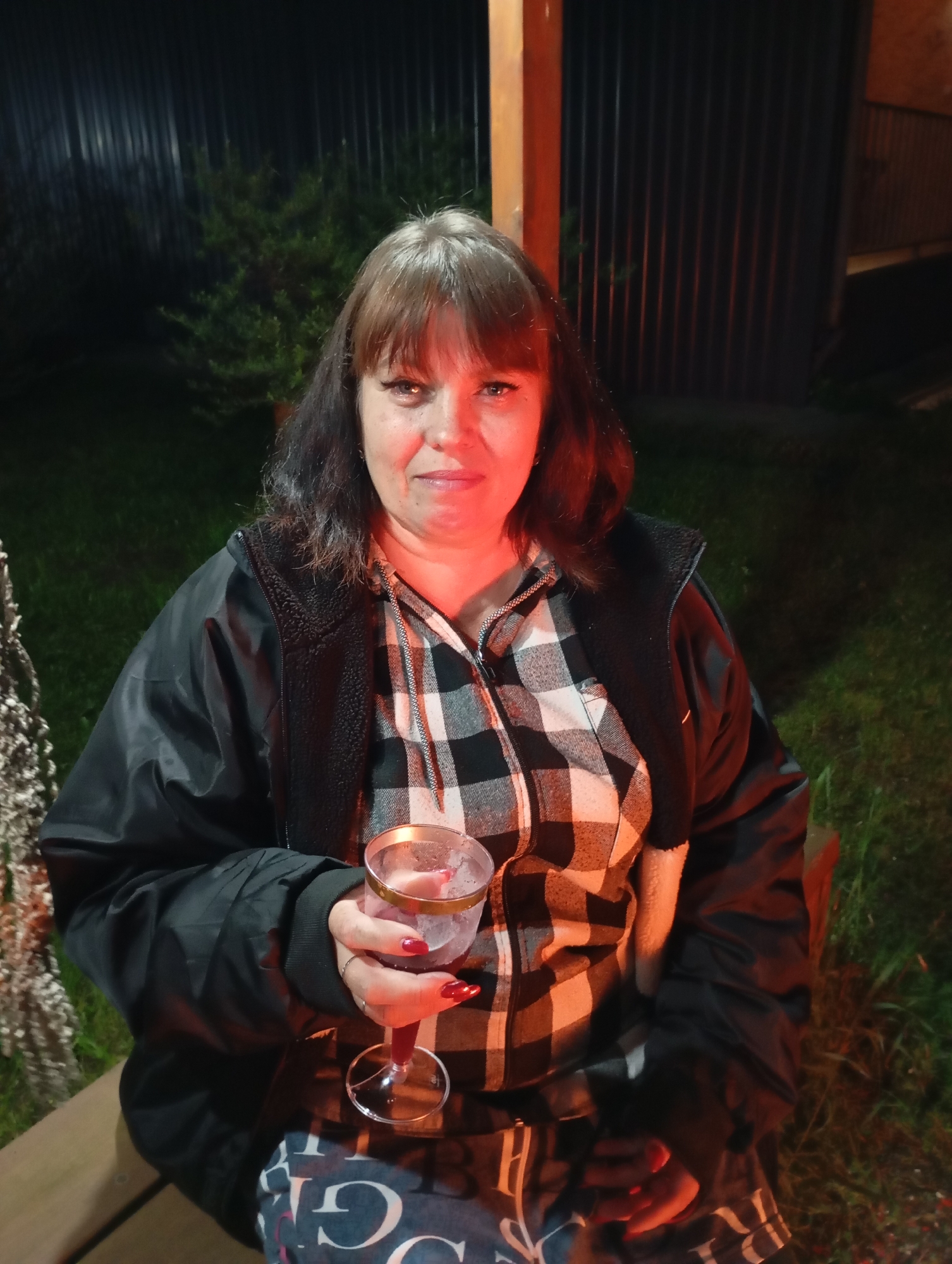 In Malyi Bukor, Russia, a woman with shoulder-length brown hair and bangs sits outdoors at night, holding a stemmed glass containing a reddish liquid. She is wearing a black jacket over a black and white plaid shirt. Her fingernails are painted red. The background features a dark, corrugated metal fence and some greenery, illuminated by red light. The woman is seated on a wooden surface, and the ground is covered in grass.
