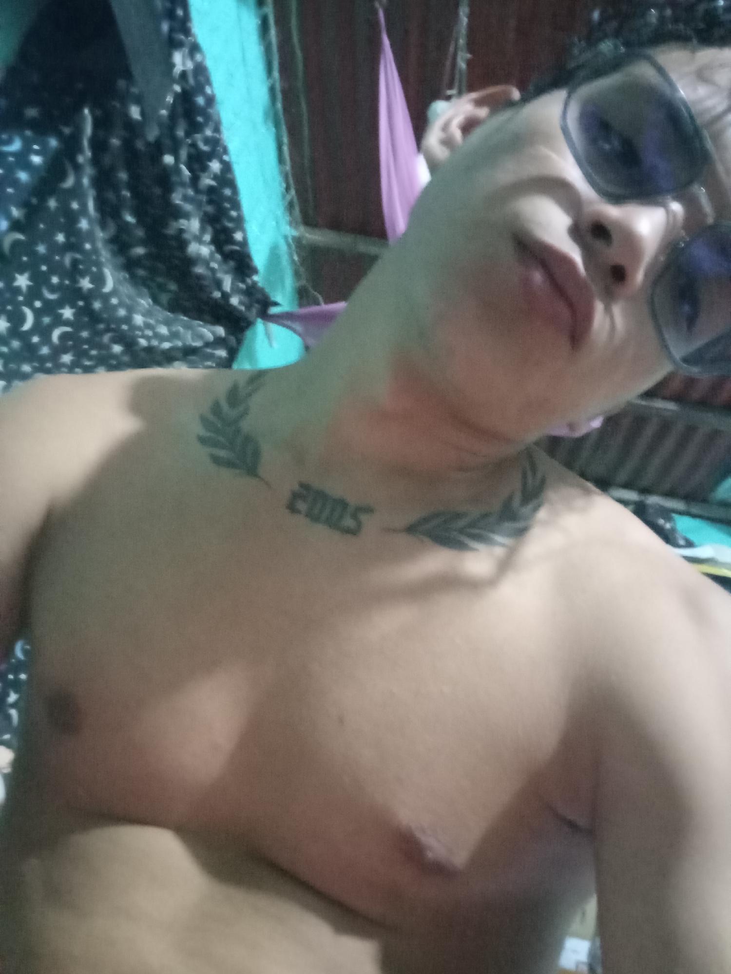The image is a close-up, angled selfie of an adult male, taken indoors in Altagracia, Venezuela. The individual is shirtless, exposing his bare chest, shoulders, and neck. He has light skin and is wearing dark-lensed sunglasses, obscuring his eyes. His head is tilted to the viewer's left, and his lips are slightly pursed in a pouting expression. On his upper chest/collarbone area, a prominent tattoo displays the numbers "2005" centrally, flanked by two symmetrical dark-inked designs resembling olive or laurel branches, one extending towards each shoulder.
The background is an indoor, somewhat cluttered domestic environment. To the viewer's left, a dark fabric with a repeating pattern of white stars and crescent moons is visible. A section of light blue/teal fabric or wall is present in the upper left. Behind the subject's head, a piece of purple fabric, possibly part of a hammock or curtain, hangs. Dark wood paneling or a wall surface is visible in the upper right background. Indistinct metallic or rope elements are also partially visible hanging from above in the top center. The scene is illuminated by ambient indoor light, casting soft shadows.