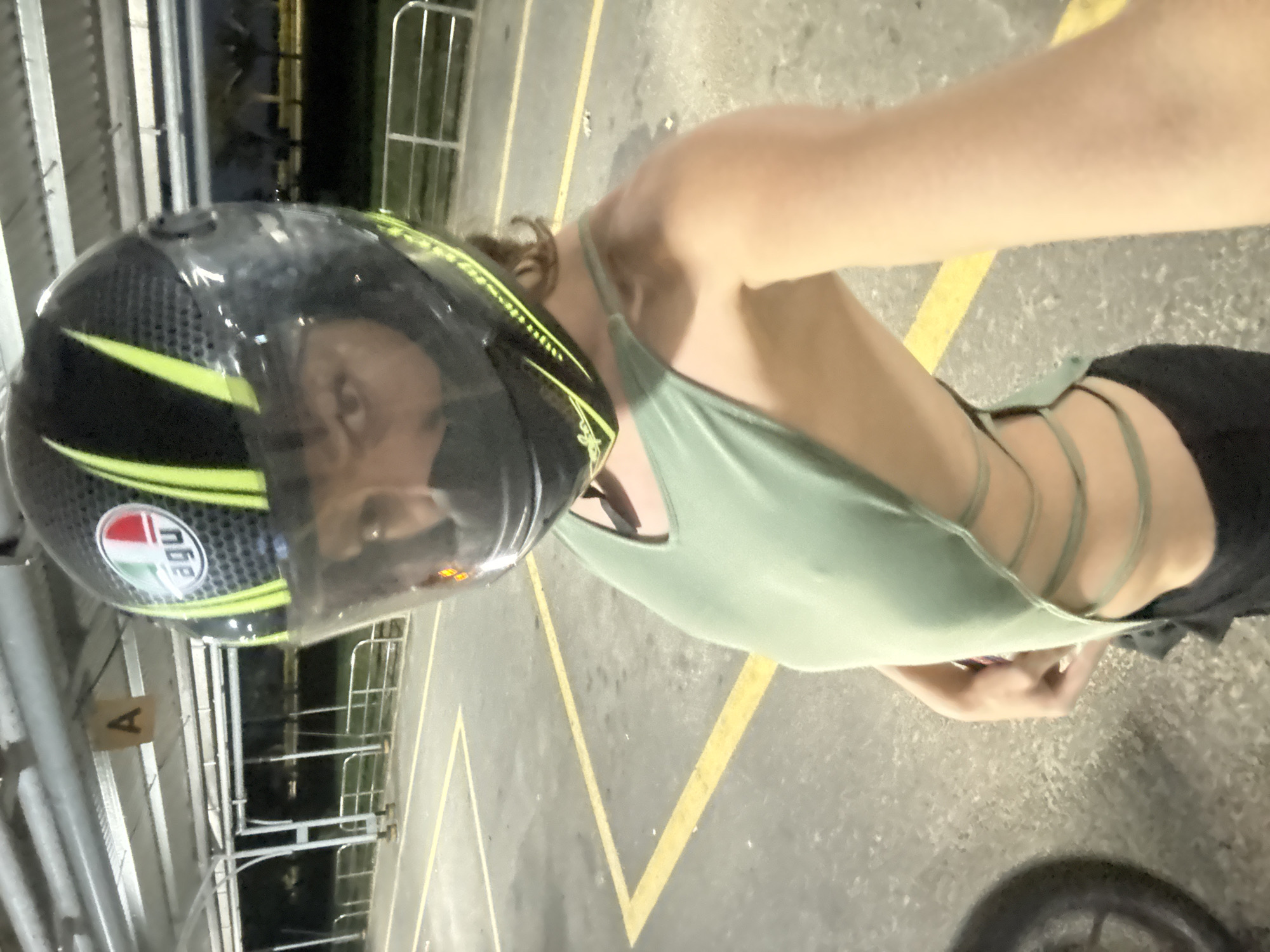 A person wearing a motorcycle helmet and a light green halter top is standing in what appears to be a parking area. The ground is concrete with yellow parking space markings. The helmet is black with yellow and white accents, and the person's face is visible through the helmet's clear visor. The halter top has multiple straps that cross in the back. The background shows what seems to be the entrance to a parking garage. The specific location within Đà Nẵng is not discernible from the image.