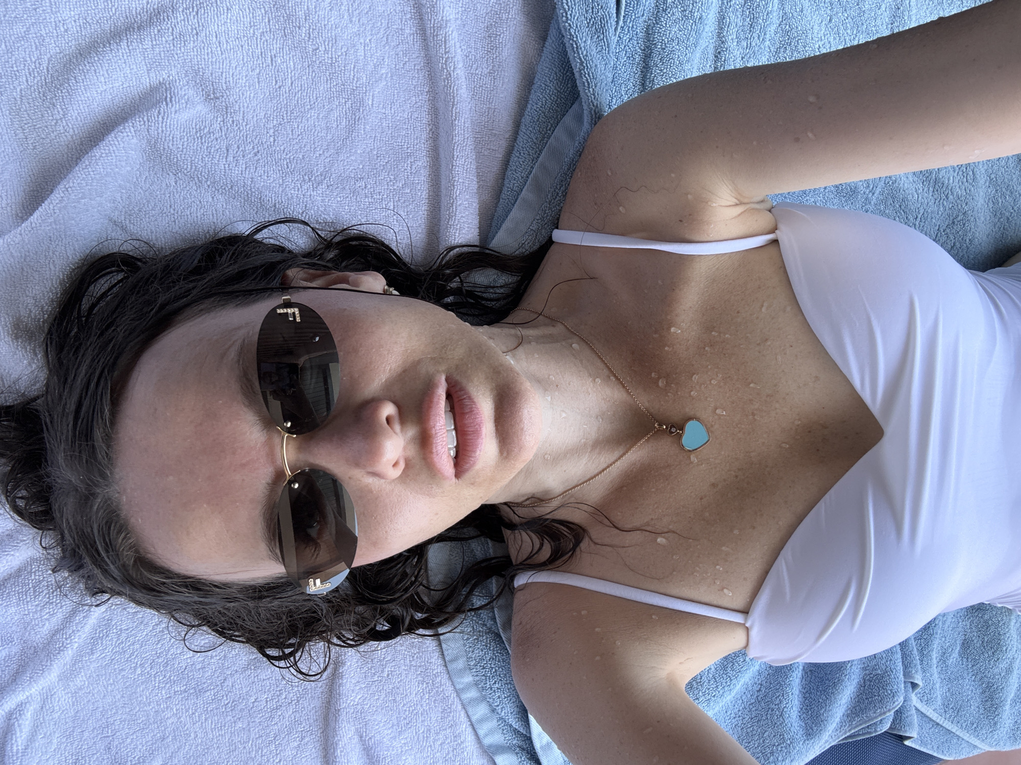 A woman lies on her back on a blue towel, her head to the left and her body extending towards the right. She wears a white swimsuit and sunglasses, with her dark hair spread out around her head. Water droplets are visible on her skin, suggesting she is either wet or has been exposed to humidity. A delicate gold chain with a turquoise heart-shaped pendant rests on her chest. The background consists of a white towel, also with a slight texture. The overall impression is one of relaxation, possibly in an outdoor setting given the presence of water droplets. The location within Dubai, United Arab Emirates is not discernible from the image itself.