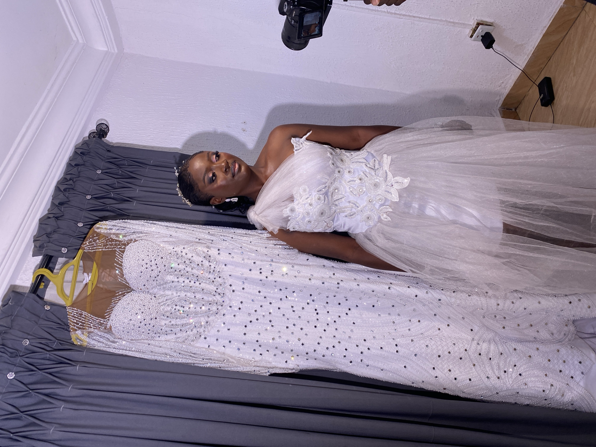 In a room with white walls and a wooden floor, a young Black woman stands to the right of a heavily beaded white wedding dress hanging on a hanger. The wedding dress is floor-length and has a sweetheart neckline with long sleeves, entirely covered in iridescent beads and sequins. The woman is wearing a short, white, one-shoulder wedding dress with a tulle skirt. She has dark skin, dark hair pulled back, and is wearing makeup and a tiara. A DSLR camera on a tripod is positioned to her right, facing her. A grey curtain with a pleated pattern hangs on the left side of the image. The scene appears to be in a residential setting in Bukuru, Nigeria.