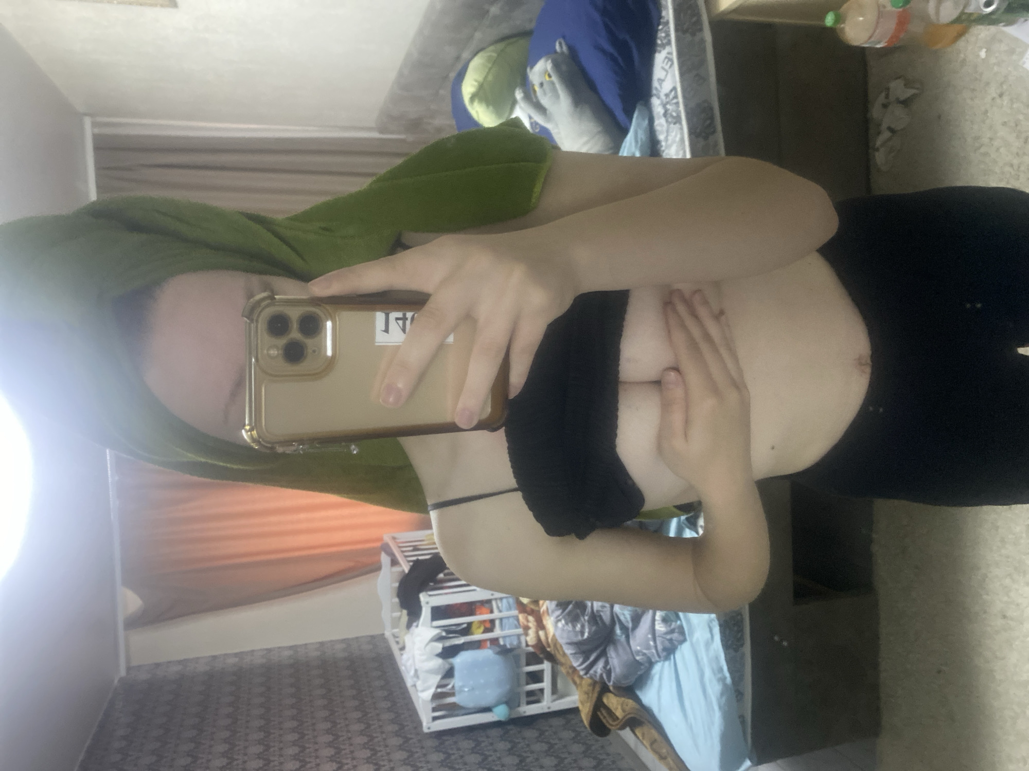 An individual is depicted in a mirror selfie taken indoors, likely a bedroom or living area in Ternopil, Ukraine. The image is oriented sideways, with the person holding an iPhone with a gold-colored case. A green towel is wrapped around their head, suggesting a recent shower or hair wash. They are wearing a black strapless top or bandeau and black bottoms, with one hand holding the phone and the other covering part of their chest.
The background reveals a domestic setting. To the left, light-colored curtains, possibly orange or brown, are visible. A white crib or playpen, containing what appears to be clothing and a light blue stuffed toy, is situated towards the bottom left. A bed with blue bedding, a blue pillow, and a grey stuffed animal lies behind the person. On a surface to the right, several bottles, including one with a green cap, and some crumpled items are visible. The floor is covered with a patterned carpet. The person's face is largely obscured by the phone, preventing the discernment of specific emotions. Text on a sticker on the phone case is partially visible and reversed, making it difficult to read accurately.