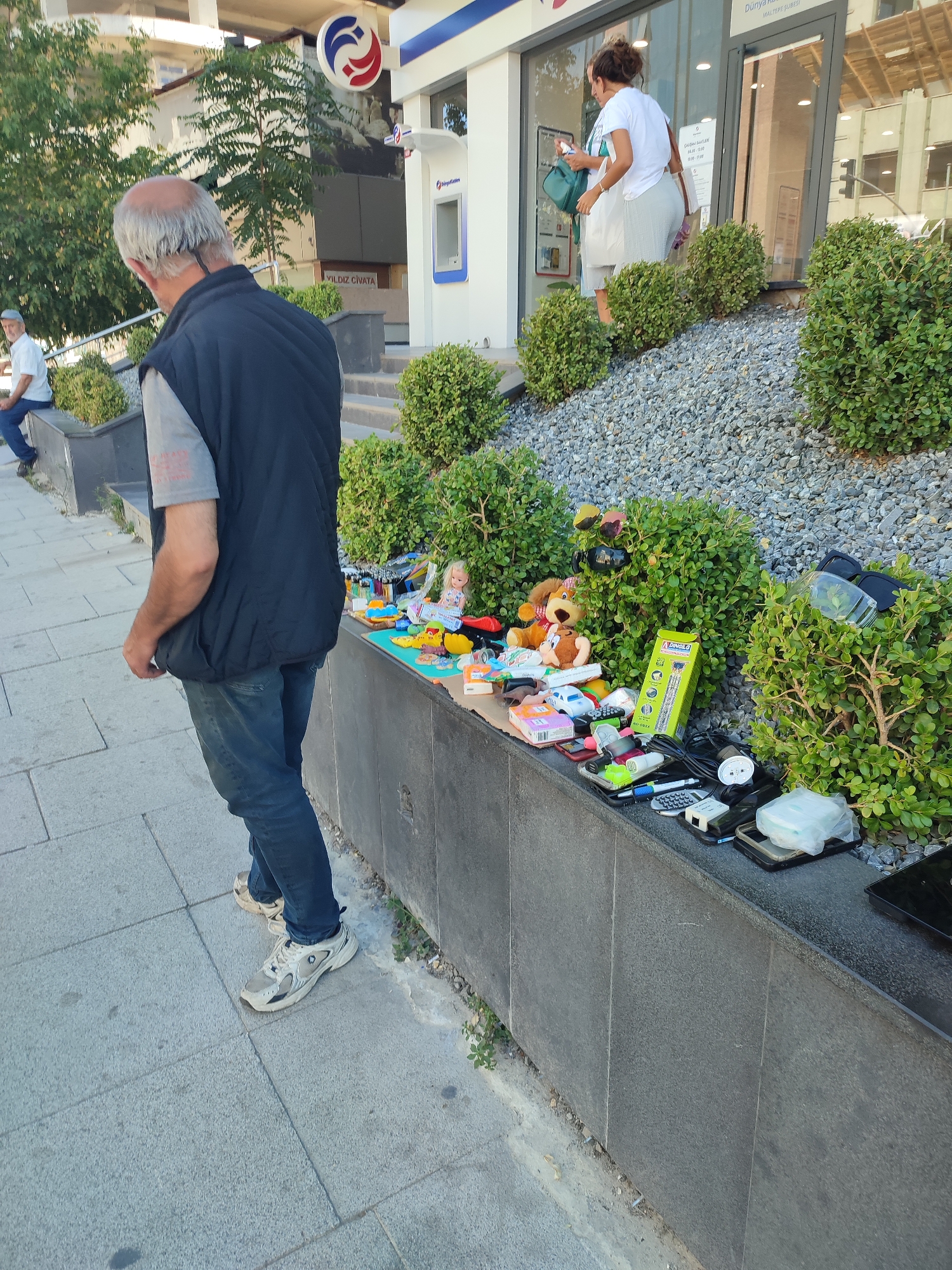 An elderly man with gray hair, wearing a dark vest over a gray t-shirt and blue jeans, stands on a paved sidewalk in Maltepe, Turkey. He is facing away from the camera, looking towards a display of items on a raised stone ledge. The ledge is lined with small, manicured green bushes. On the ledge, various objects are laid out, including stuffed animals, toy cars, electronic devices such as remote controls and cell phones, a doll, and a yellow box labeled "DANDILO". Behind the ledge, a building with large windows and a sign that reads "DÜNYA KALİTE" is visible. A woman in a white t-shirt and gray skirt is seen exiting the building. The ground surrounding the display is covered in gray gravel. The scene suggests a street vendor or a temporary sale of goods.
