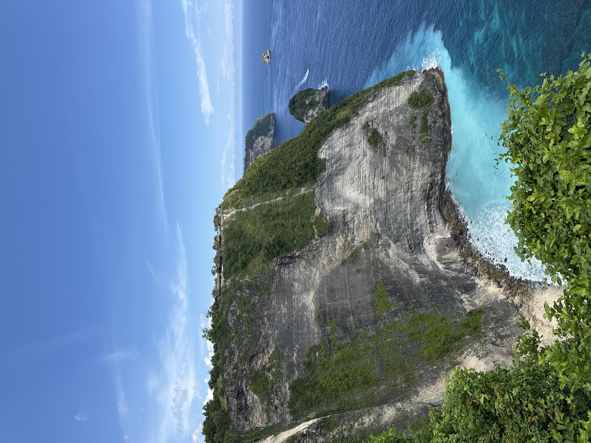 This image showcases a dramatic coastal landscape at Nusa Penida Island in Bunga Mekar, Indonesia. Dominating the scene is a towering, layered cliff face with varying shades of grey and dark rock. The upper portion of the cliff is covered in lush, green vegetation. A narrow path is visible along the cliff edge. Below the cliff, the turquoise ocean meets the rocky coastline. Two smaller, rocky islands with vegetation are situated in the deep blue sea. A boat is travelling near the islands. The sky is a clear blue with a few wispy clouds. Green foliage is prominent in the bottom right of the frame.