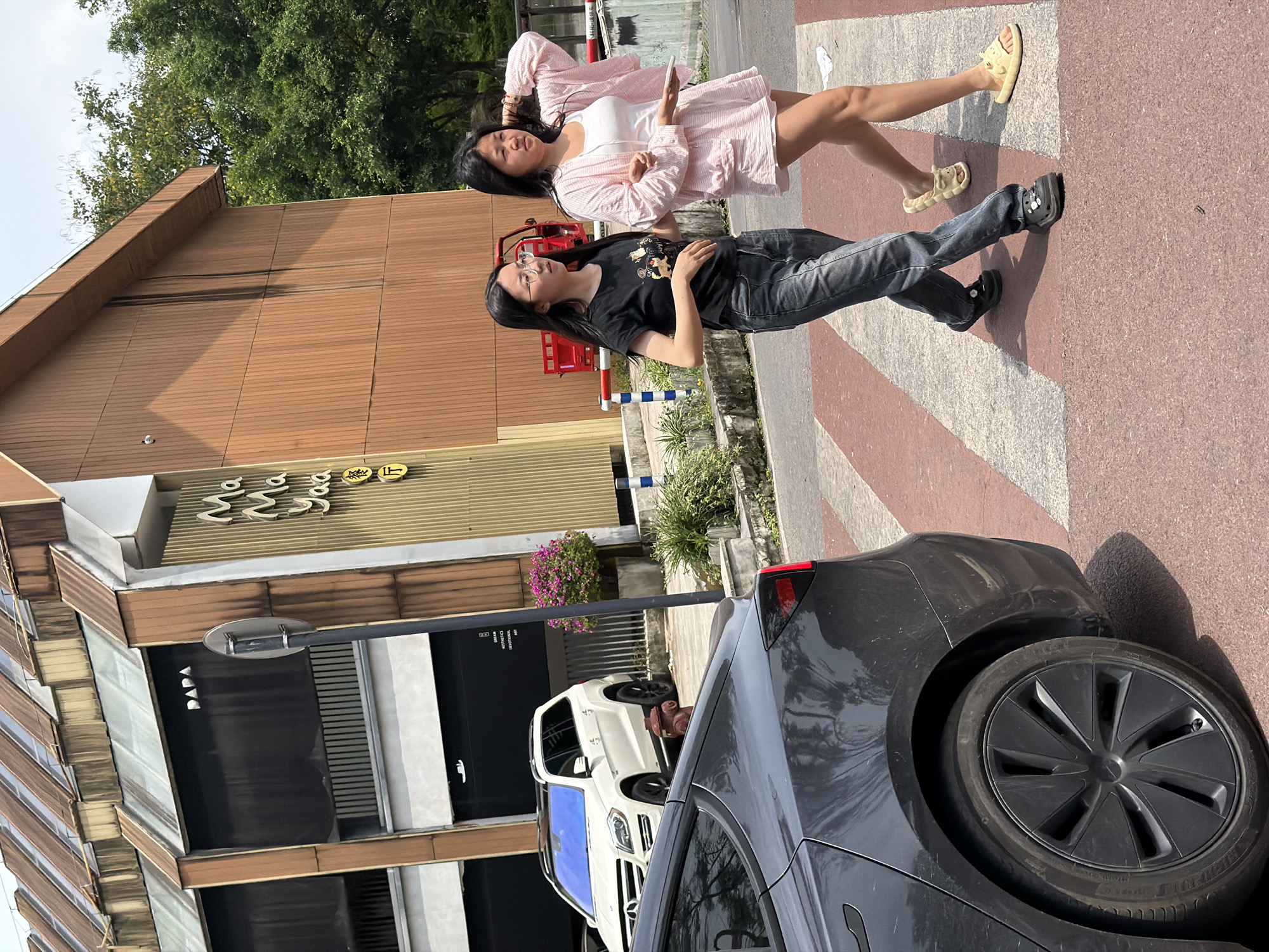 Two women stand on a red-and-white striped crosswalk in front of a building. The woman in the foreground wears a black T-shirt, jeans, and black shoes. The woman behind her wears a light pink dress and yellow sandals. A black car is parked on the right side of the frame. Across the street are two white cars. The building in front of them has "PAPA" written on a black sign and Chinese characters above. The location is identified as the Chongqing High-tech Zone, China.