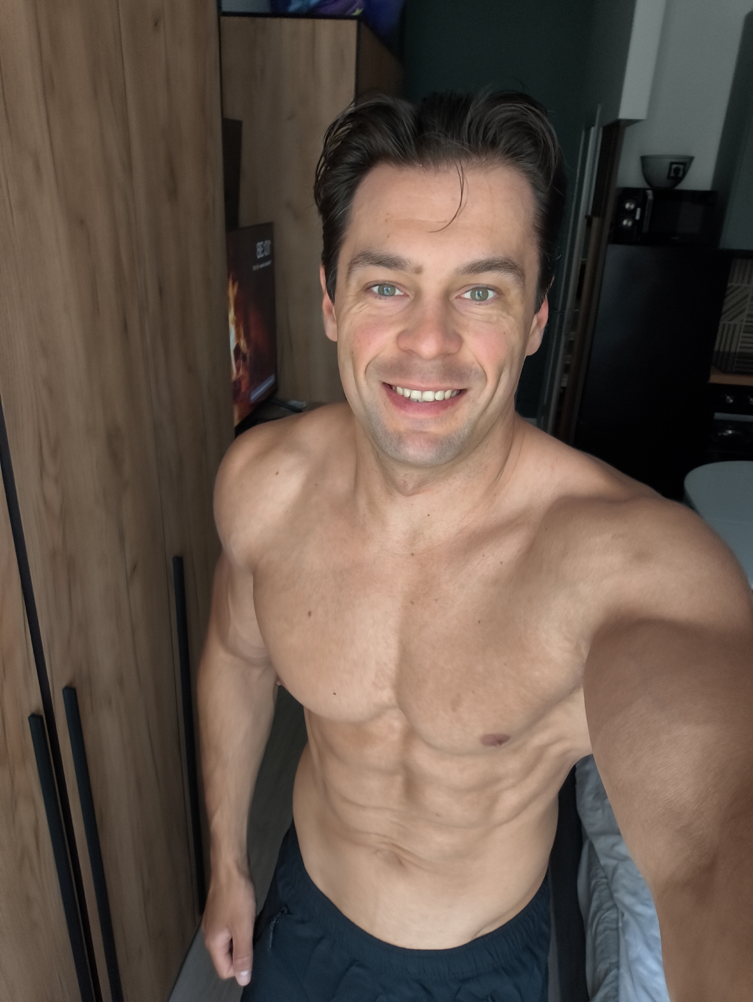 A shirtless man with a muscular physique and light brown hair smiles at the camera in an indoor setting. He is wearing dark blue shorts. Behind him, a wooden wardrobe is visible on the left, and a television screen with an image of flames is partially seen. On the right, a microwave and a bowl are on a black surface, and a bed with grey bedding is partially visible. The scene appears to be a bedroom or living area. There is no information in the image that indicates it is located in Moscow, Russia.