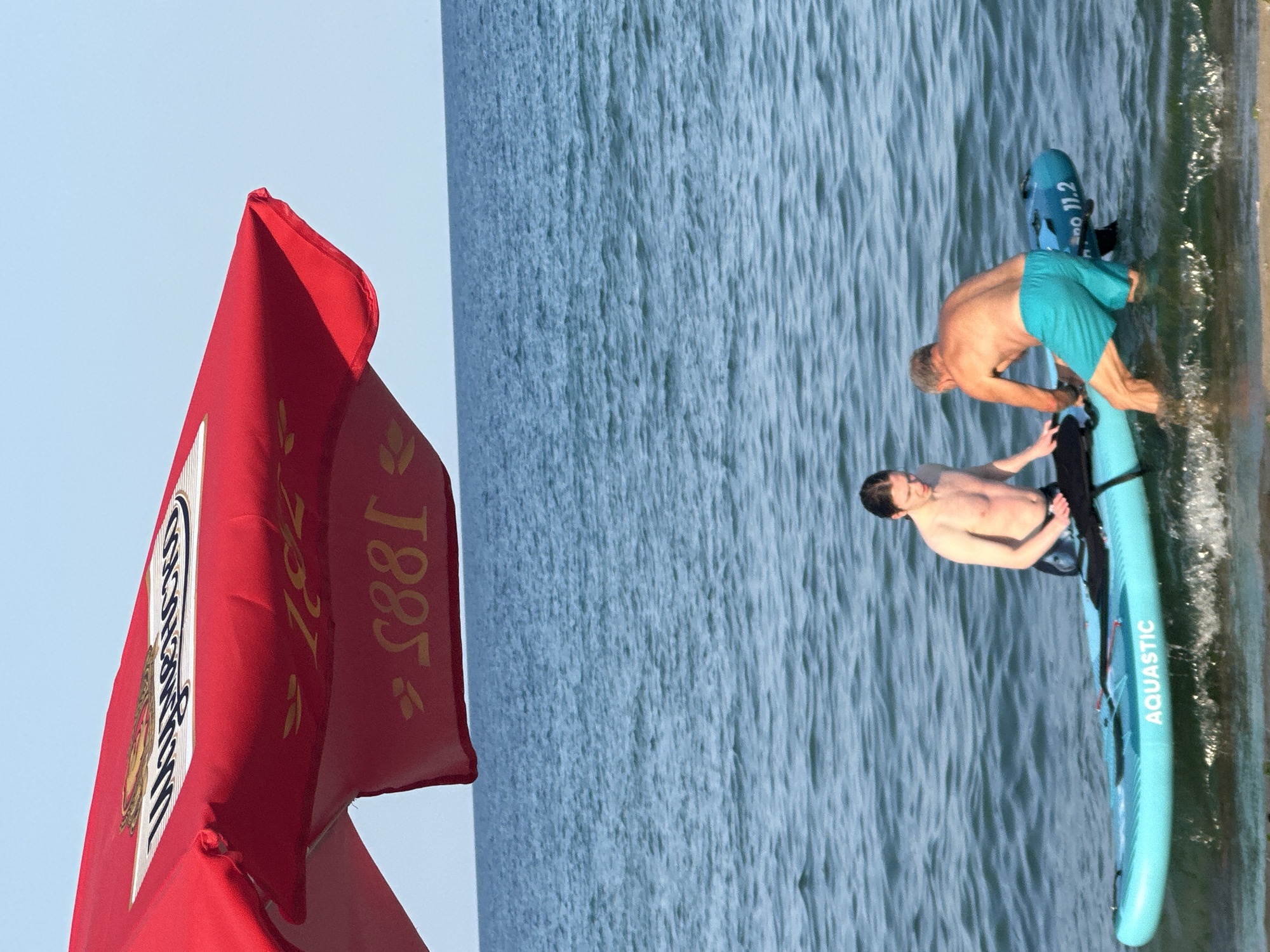 Two shirtless men are on a paddleboard in the shallow ocean water. One man, with long dark hair, is sitting on the paddleboard. The other man, with gray hair, is standing on the paddleboard, holding onto it as he enters the water. Both men are wearing swim trunks. The paddleboard is light blue and has "AQUASTIC" written on its side. A large red umbrella with gold lettering is visible in the upper left corner of the image, partially obscuring the clear blue sky. The water is calm with small waves.