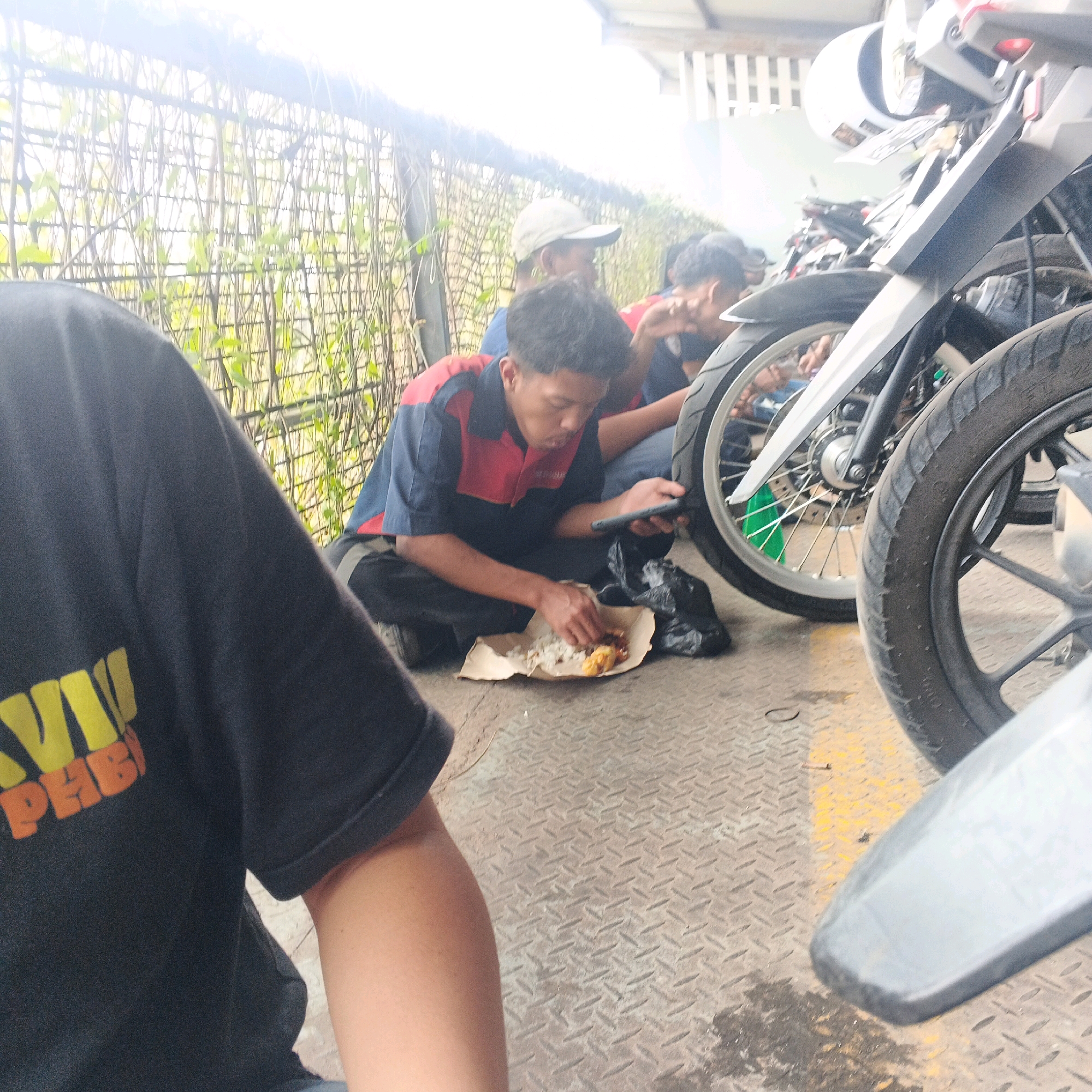 In Banjararum, Indonesia, a group of young men are seated on a diamond plate metal floor. One man in a blue and red uniform is eating from a paper plate, holding a mobile phone in his left hand. Another man in a beige cap and a red and blue shirt is partially visible behind him. The foreground shows the arm and shoulder of a person wearing a black t-shirt with yellow and orange lettering. Several motorcycles are parked closely together, with their wheels and parts prominently featured in the image, particularly the front wheel of a white and silver motorcycle. The background consists of a metal fence with green foliage growing through it, and a building with a slatted roof. The lighting suggests it is daytime, possibly overcast.