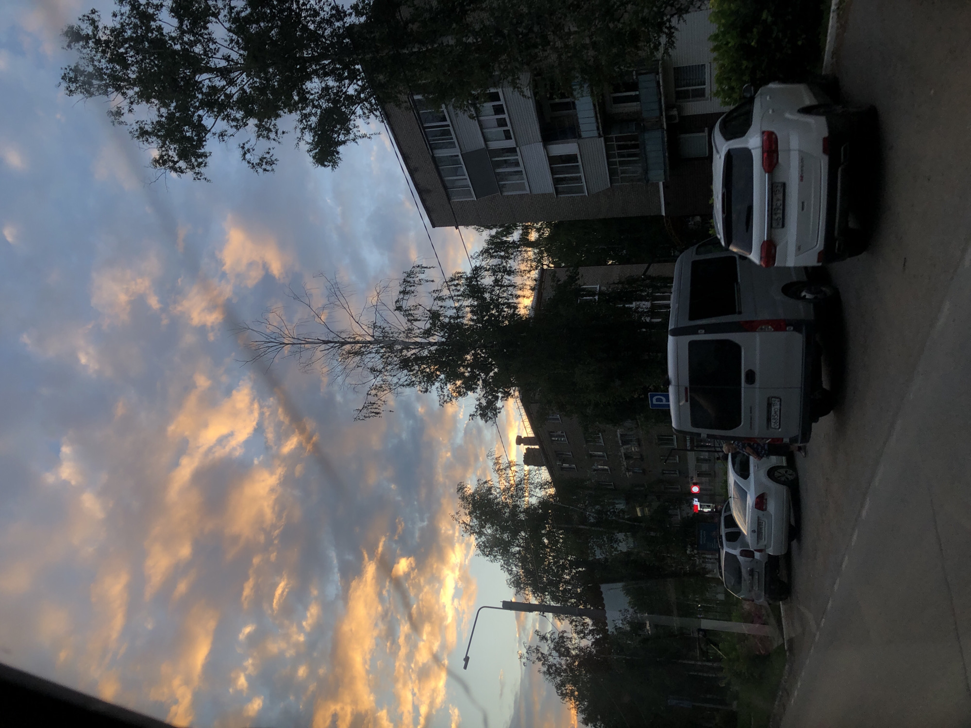 The image depicts a residential street in Chaikovsky, Russia, during sunset. The sky is filled with dramatic clouds painted in shades of orange, pink, and blue, indicating the sun is low on the horizon. Several apartment buildings, typical of Soviet-era architecture, line the street, their facades appearing somewhat aged. In the foreground, a line of parked cars is visible, including a white SUV, a silver van, a white sedan, and a dark SUV. The street is paved, and a sidewalk with some greenery can be seen. A lamppost stands on the left side of the frame. The overall atmosphere is calm and serene, characteristic of an evening in a residential area.