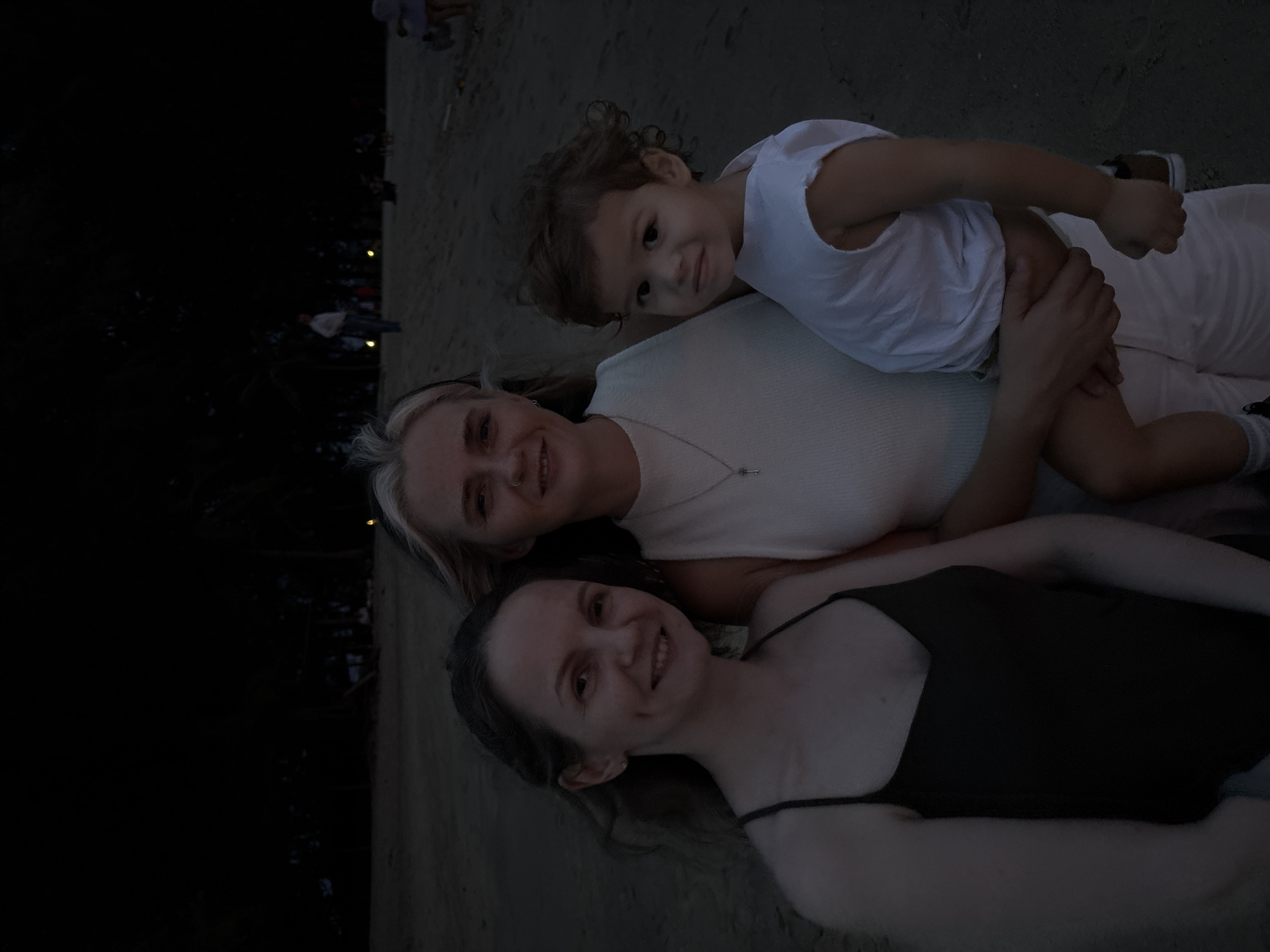 In the city of Choeng Thale, Thailand, three individuals are captured in a low-light photograph on a sandy beach at dusk. A woman on the left, with long dark hair, wears a dark-colored tank top and smiles at the camera. Beside her, a woman with blonde hair, wearing a white ribbed top, holds a young child. The child, a boy with curly brown hair, is dressed in a white t-shirt and is being held by the woman. He looks towards the camera with a neutral expression. In the background, palm trees are visible, silhouetted against the darkening sky, and faint lights suggest other people are present further down the beach. The ground is composed of sand, characteristic of a beach environment.