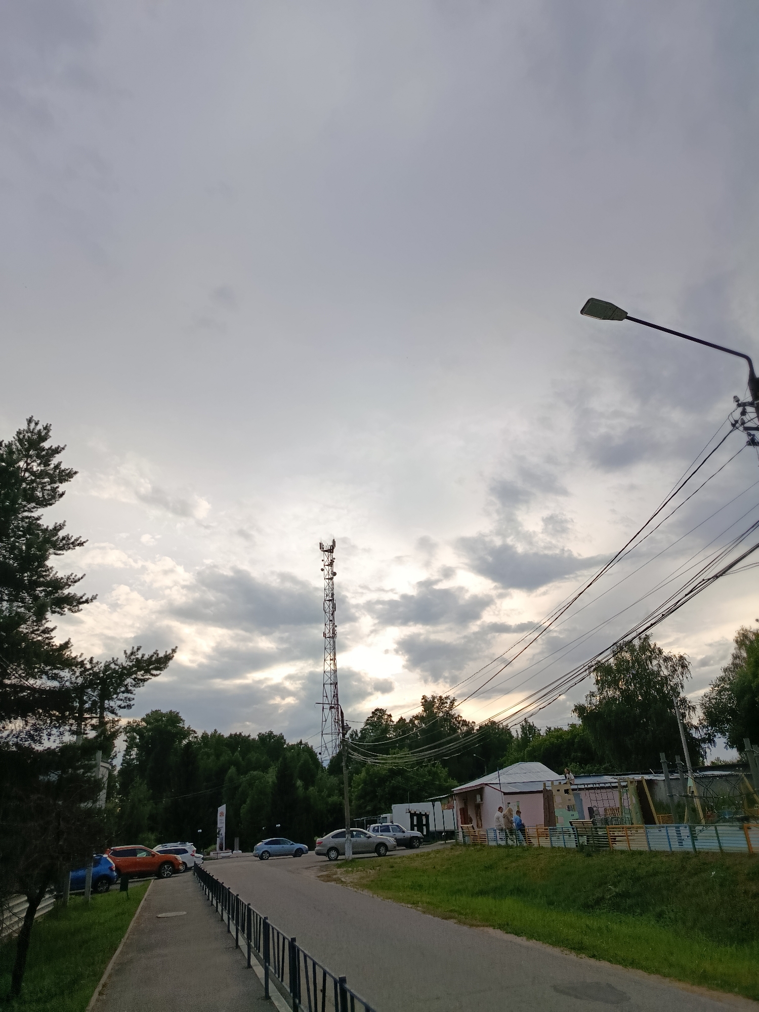 The image depicts a view of a telecommunications tower and a street in the city of Vereya, Russia. The sky is overcast with clouds. A tall, lattice-like telecommunications tower stands prominently in the background. To the right of the tower, a street stretches into the distance, bordered by a sidewalk and a grassy verge with a black metal fence. Several parked cars are visible along the side of the road, including an orange SUV, a white car, and a blue sedan. A low building with a light-colored roof and pink walls is situated to the right of the street, with a playground area behind it. Several individuals are standing near the building. Power lines are strung across the upper portion of the image, supported by poles. A streetlamp is also visible on the right side. On the left side of the image, the branches of a large evergreen tree extend into the frame. The scene appears to be a public area, possibly a residential or community space.
