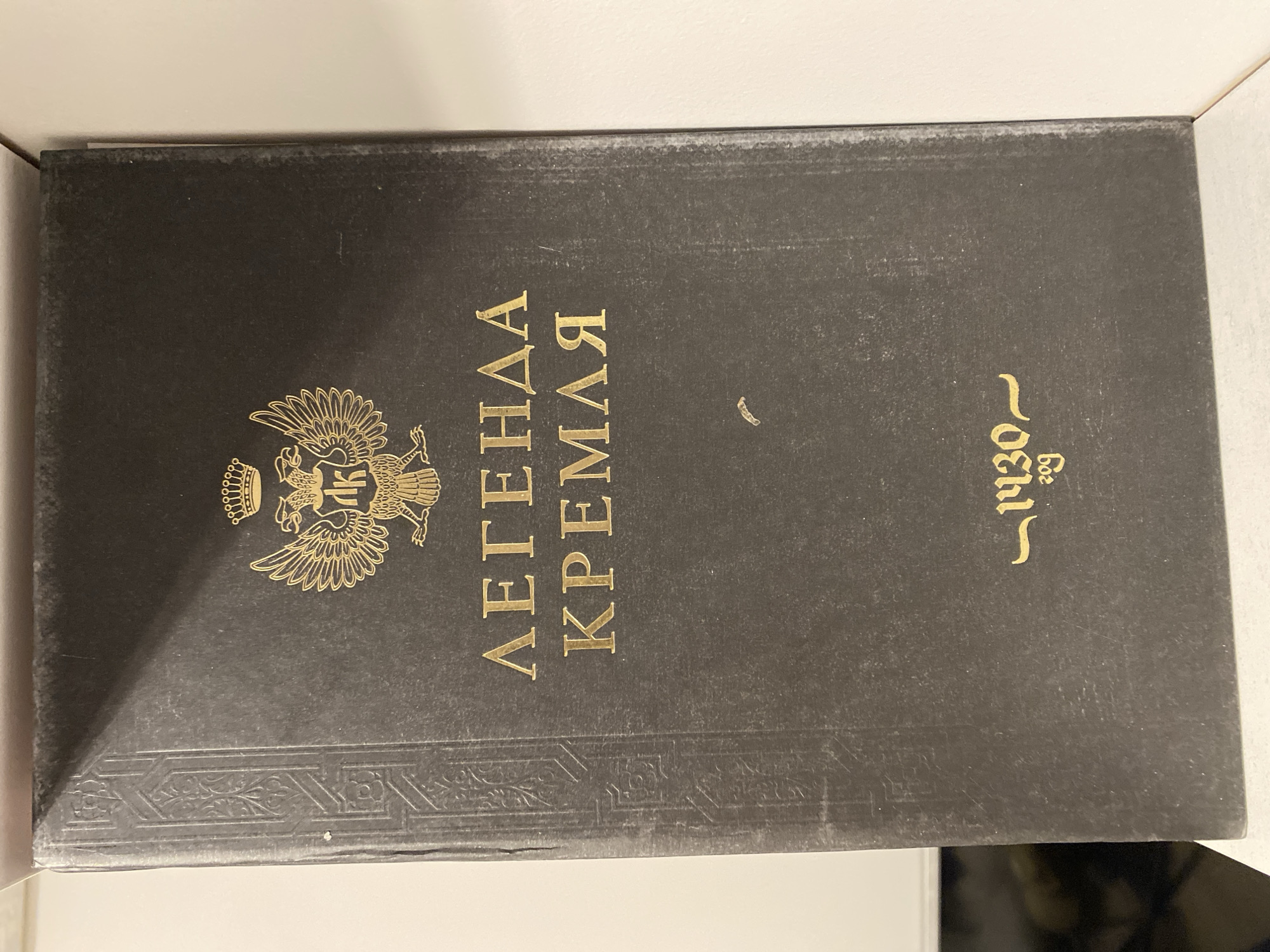 A close-up studio shot depicts a dark-covered book, oriented with its spine facing the viewer. The spine features a gold-colored emblem of the Russian double-headed eagle with a crown above it, along with the words "ЛЕГЕНДА КРЕМЛЯ" (LEGEND OF THE KREMLIN) in Cyrillic. To the right of the text, the numbers "1930" are also printed in gold. The spine shows signs of wear and age, including scuff marks and slight fading. The location of the book is not specified.