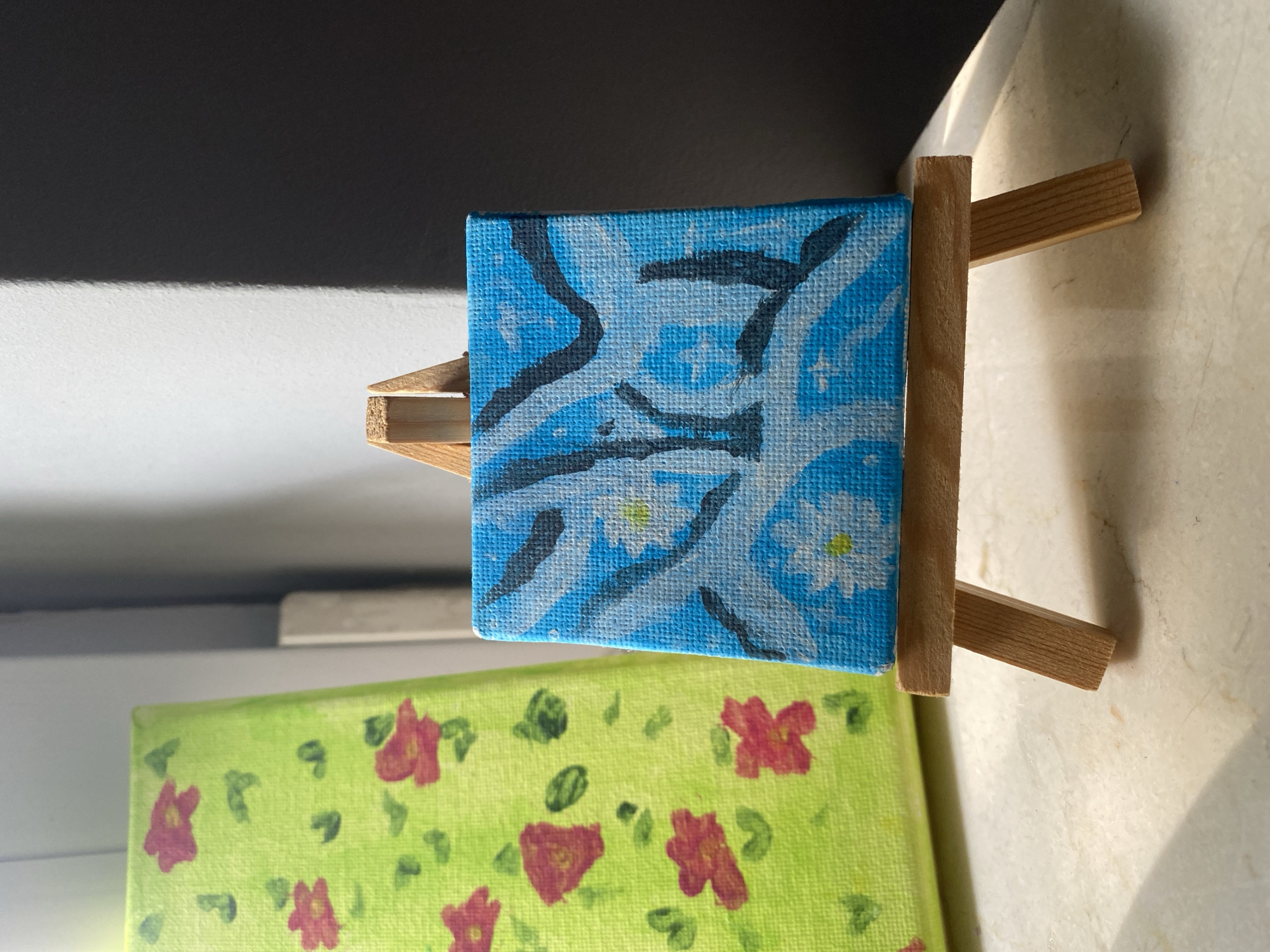 The image shows two small canvas paintings. The painting in the foreground is square, with a blue background and abstract dark blue and white shapes, possibly representing stylized flowers or patterns. It is displayed on a miniature wooden easel. Behind it, to the left, is another rectangular canvas, mostly out of focus, painted with a bright green background and scattered red flowers and green leaves. The scene is lit by natural light, casting shadows on the wall behind the easel. The surface the easel rests on appears to be a marble or stone countertop. The setting is indoors, with a window or light source to the right casting a shadow. There are no people or animals visible in the image. There is no information in the image to suggest a specific location in the city of Łódź, Poland.