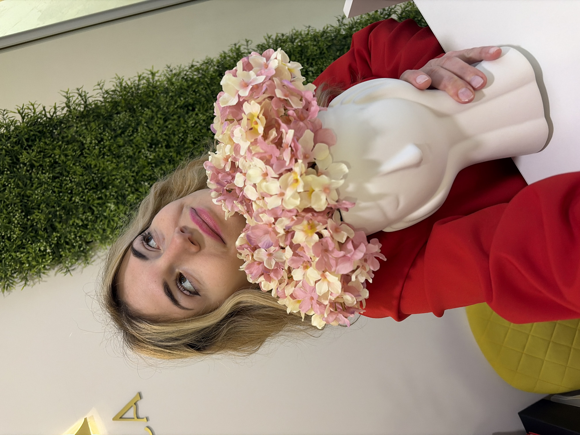 A woman with blond hair is holding a white, head-shaped vase filled with pink and white flowers. She is wearing a red top. The background includes a wall with faux greenery, a white wall, and a yellow stool. The location is Budva, Montenegro.