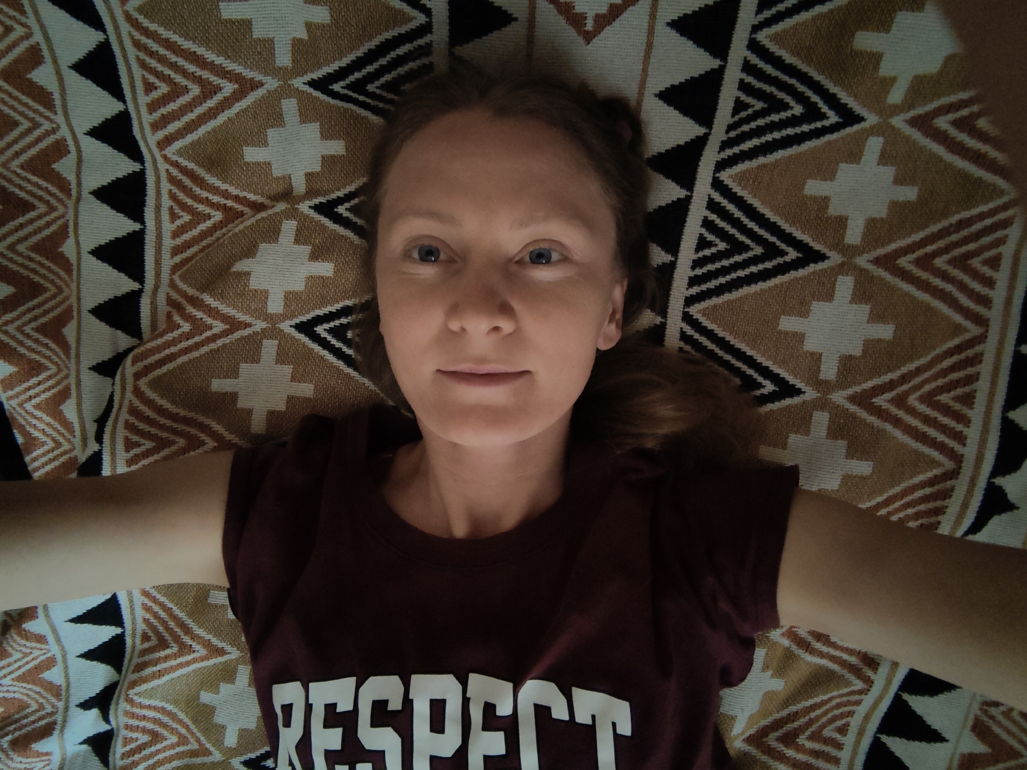 The image shows a young woman lying on a patterned blanket. She is wearing a maroon t-shirt with the word "RESPECT" printed in white letters across the chest. Her arms are outstretched to the sides, and her head is slightly tilted back. The blanket features a geometric pattern with triangles and diamond shapes in shades of brown, white, and black. The woman's hair is light brown and appears to be pulled back. Her eyes are blue and she is looking directly at the camera. The scene is well-lit, with natural light illuminating her face. The location is described as Зуйское сельское поселение, Ukraine.