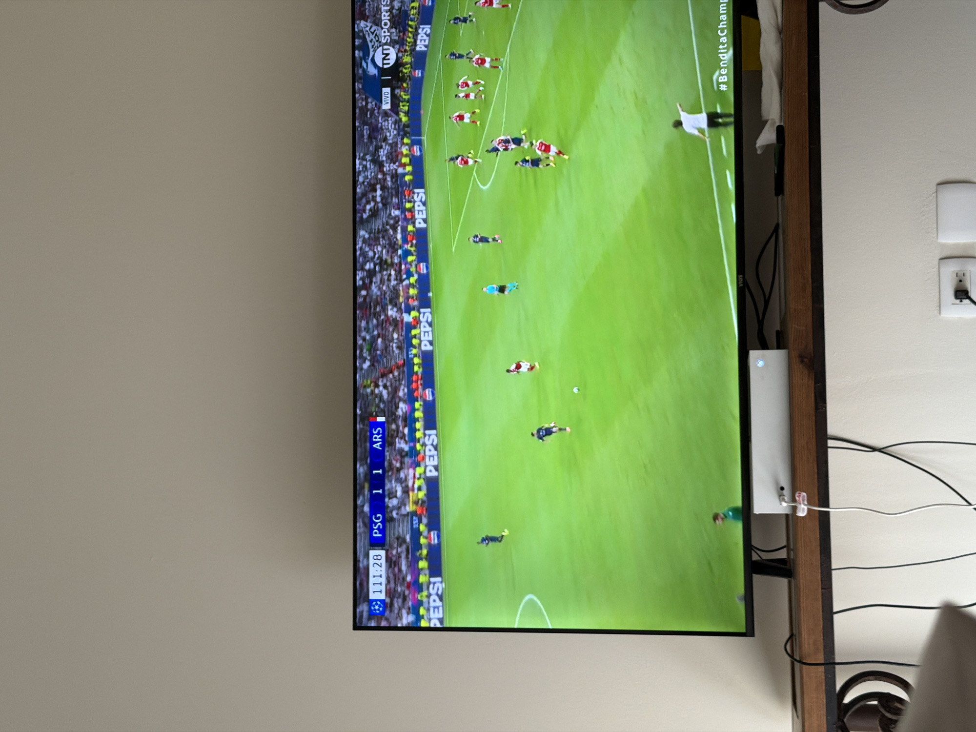 A television is displaying a soccer match with PSG playing Arsenal. The score is tied 1-1 with the game clock at 111:28. The television is hanging on the wall of what appears to be a residential home, located in Cancun, Mexico, close to electrical outlets.