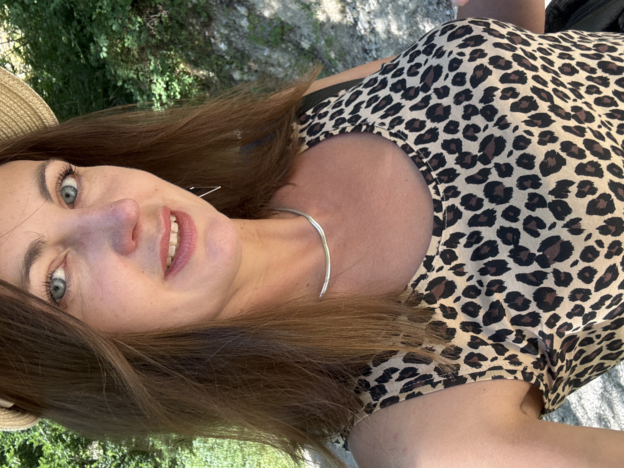 An outdoor, medium shot captures a woman in a close-up, selfie-style photograph. She is wearing a straw hat and a leopard print tank top, with a silver choker necklace adorning her neck. Her brown hair is parted in the center and falls past her shoulders. Her blue eyes are looking towards the camera, and her lips are parted in a slight smile, revealing her teeth. The background is a blurred natural environment with green foliage and a gravelly path, suggesting an outdoor setting that could be found on the outskirts of Saint-Claude, France.