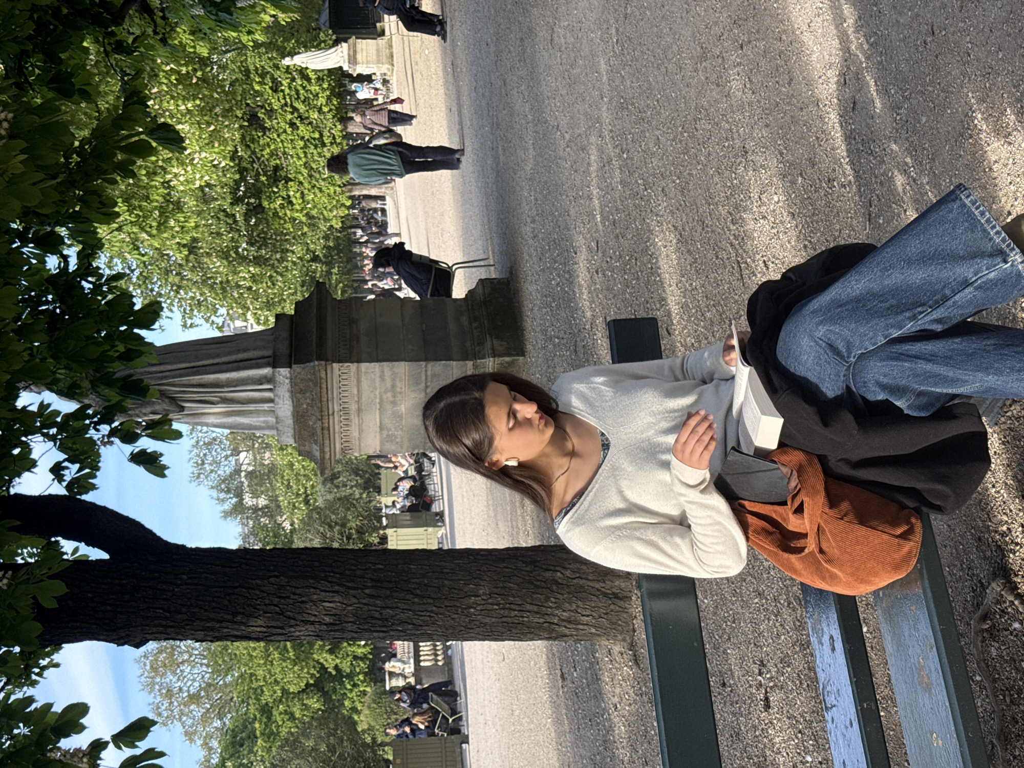 The photograph shows a young woman sitting on a park bench. The location is likely the Jardin du Luxembourg in Paris, France, given the visible statue and layout of the park. She is wearing a white sweater and jeans, and a black coat and brown corduroy bag are next to her on the bench. She seems to be reading a book or using an electronic device. The bench is positioned along a path, with trees and foliage visible in the background. In the distance, other people can be seen walking or sitting in the park. The time of day appears to be late afternoon, with the sun casting shadows on the ground.