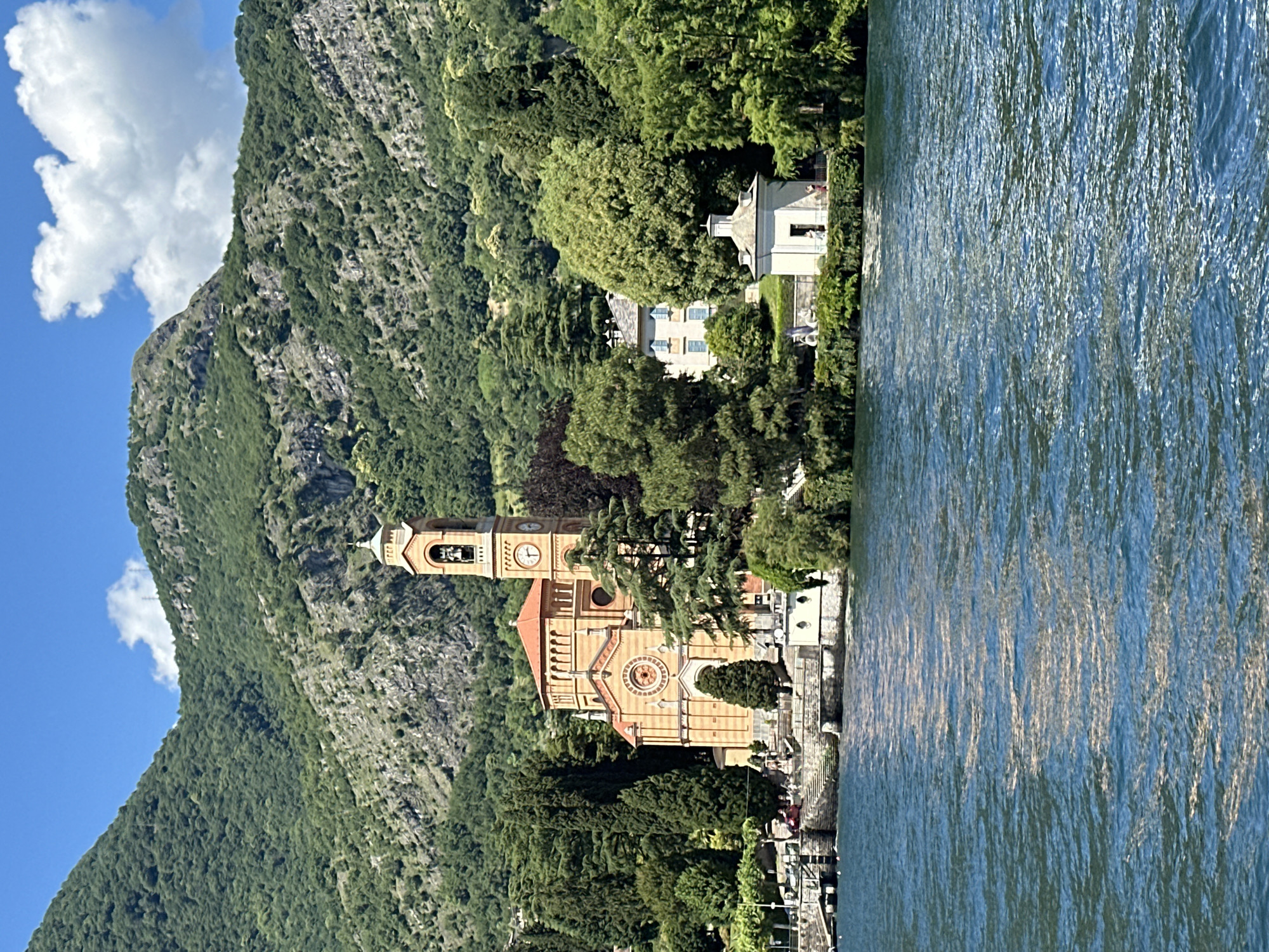 This vibrant landscape photograph captures a picturesque scene in Tremezzina, Italy, featuring a large lake, a towering mountain, and architectural elements under a bright sky.
In the foreground, the serene, blue-green waters of the lake stretch across the bottom of the frame, showing gentle ripples and reflections of the sunny sky.
Dominating the midground is a beautiful, light orange-colored church, distinguished by its classical architecture. It features a prominent bell tower with a visible clock face and a domed top. A large rose window adorns its main facade. The church is situated very close to the water's edge, nestled among lush green trees.
Behind the church and extending up the left side of the frame is a steep, verdant mountain. Its rocky slopes are heavily covered with dense, dark green foliage, creating a dramatic natural backdrop. Several light-colored buildings, possibly villas or homes, are nestled among the trees on the lower slopes of the mountain; one house displays visible blue shutters.
The sky in the upper portion is a clear, brilliant blue, punctuated by a few fluffy white clouds. The overall impression is one of peaceful natural beauty combined with charming European architecture, under bright daylight conditions. There is no specific human activity visible, and no discernible text on the clock face or elsewhere.
