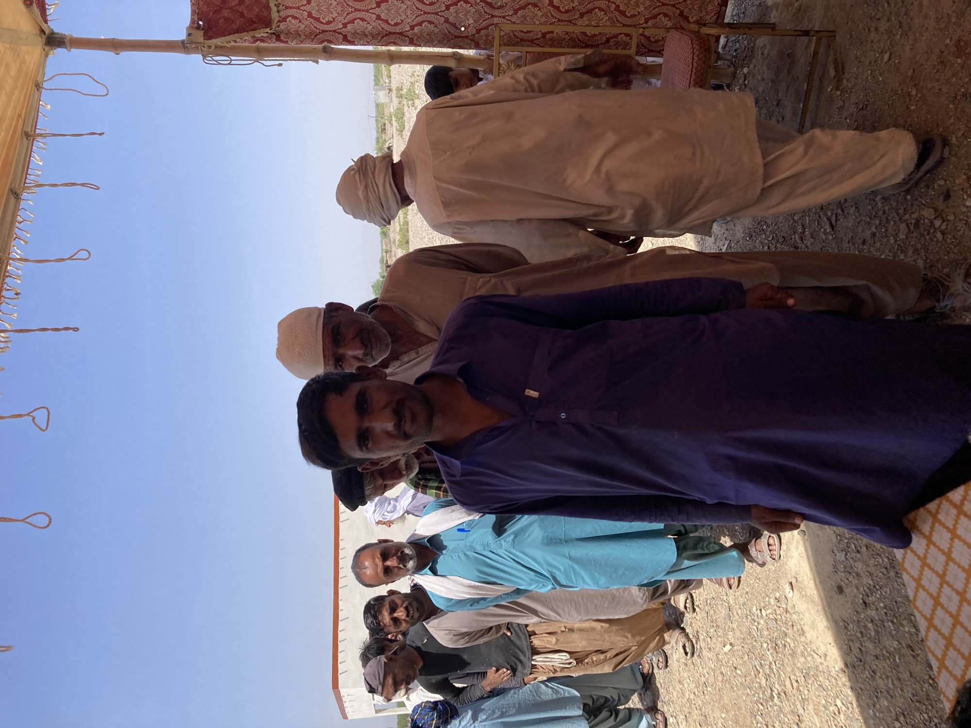 A group of men are standing together in an outdoor setting in Hussenāni, Pakistan. The men are wearing traditional Pakistani clothing, including shalwar kameez and turbans. The environment appears to be a rural area, with a dirt ground and a makeshift tent-like structure overhead.