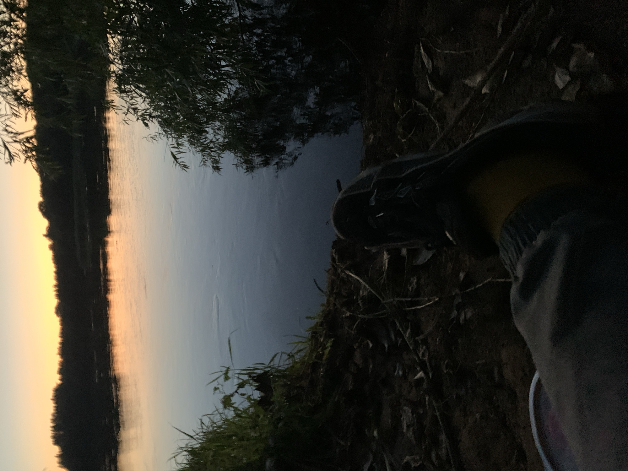 The image depicts a serene evening scene by a body of water, possibly a river, at dusk. The sky is painted with soft orange and yellow hues from the setting sun, casting a warm reflection on the water's surface. Silhouetted trees line the distant bank. In the foreground, a person's foot in a dark shoe and part of their leg in grey pants are visible, suggesting they are sitting or resting on the bank. Lush green grass grows along the edge of the water. The overall impression is one of tranquility and natural beauty during twilight. The location is the city of Kirov, Russia.